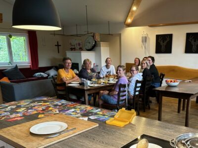 Mehrere Erwachsene sitzen an einem langen Tisch in einem gemütlichen Raum und unterhalten sich. Der Tisch ist gedeckt mit Tellern, Tassen und einem Frühstücksbuffet.