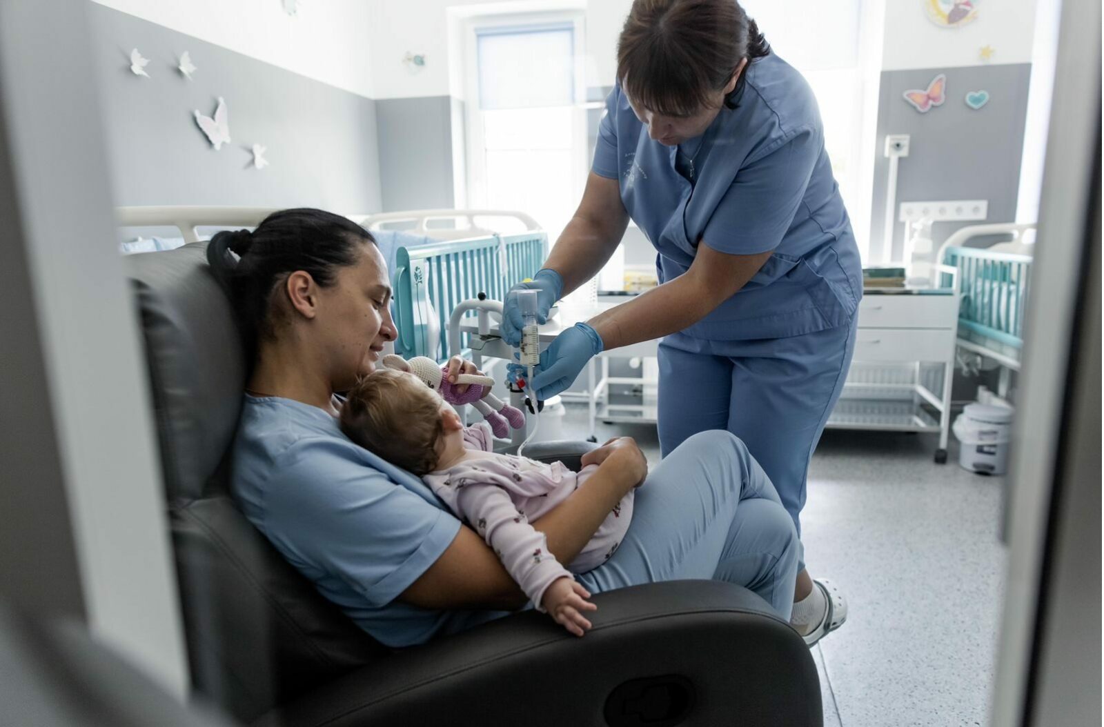 Eine Krankenschwester sitzt auf einem Sessel in einem Krankenzimmer und hält ein kleines Kind im Arm. Eine andere Krankenschwester spritzt dem kleinen Kind ein Medikament über eine Infusion. Die Krankenschwester mit dem Kind im Arm hält ein Kuscheltier hoch, um das Kind abzulenken.