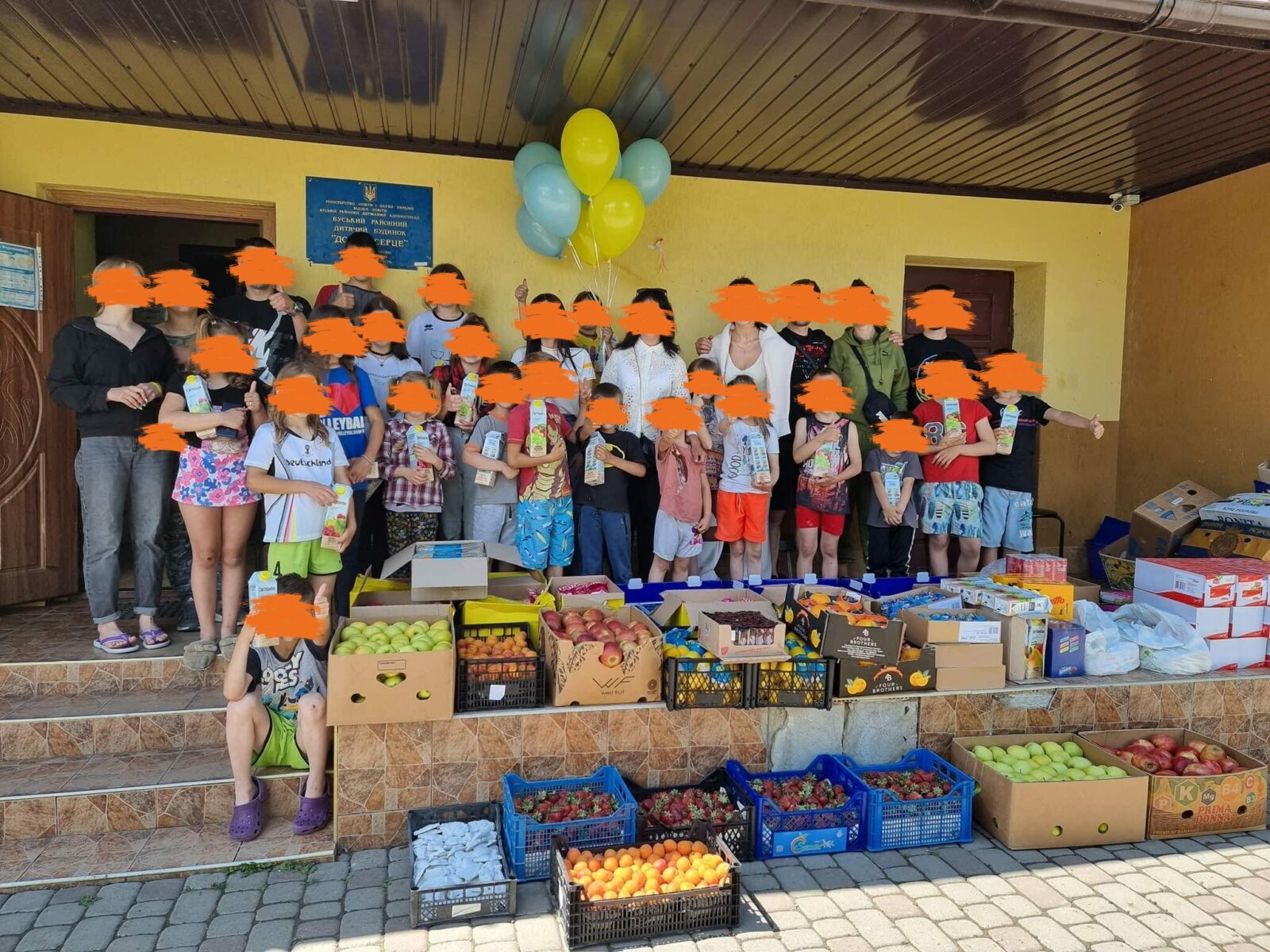 Eine große Gruppe von Kindern posiert für ein Foto vor einem gelben Gebäude. Zwei Erwachsene stehen daneben und lächeln.
