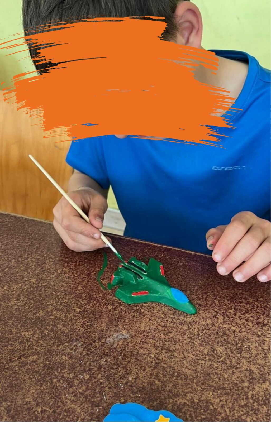 Ein Kind hält einen Pinsel in der Hand und bemalt eine grüne Figur. Der Hintergrund zeigt eine gelbe Wand.