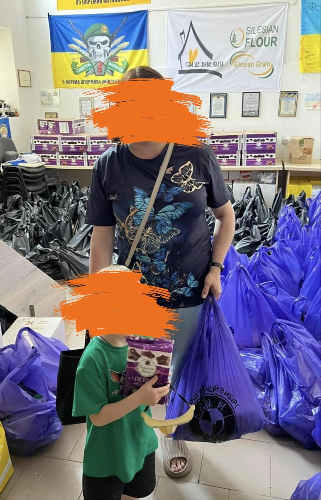 Eine Frau und ein Kin stehen in einem Raum mit mehreren Tüten voller Spenden. Die Frau hält einer dieser Tüten in der Hand und das Kind hält eine Packung Kekse in der Hand.