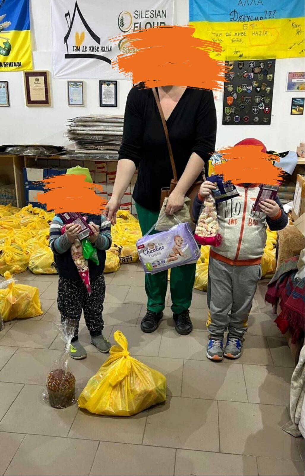 Eine Frau und zwei Kinder stehen in einem Raum voller Tüten mit Spenden. Die Frau hält eine Packung Windeln in der Hand und die Kinder halten Süßigkeiten in den Händen.