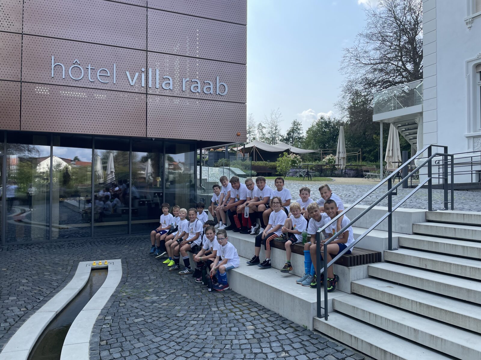 Eine große Gruppe von Kindern und Erwachsenen sitzt auf einer breiten Treppe vor einem modernen Hotelgebäude mit dem Namen „Hotel Villa Raab“. Sie tragen weiße T-Shirts. Die Kinder schauen lachend in die Kamera. Das Gebäude wirkt modern und gepflegt, mit großen Glasfenstern und einem schattigen Eingangsbereich.