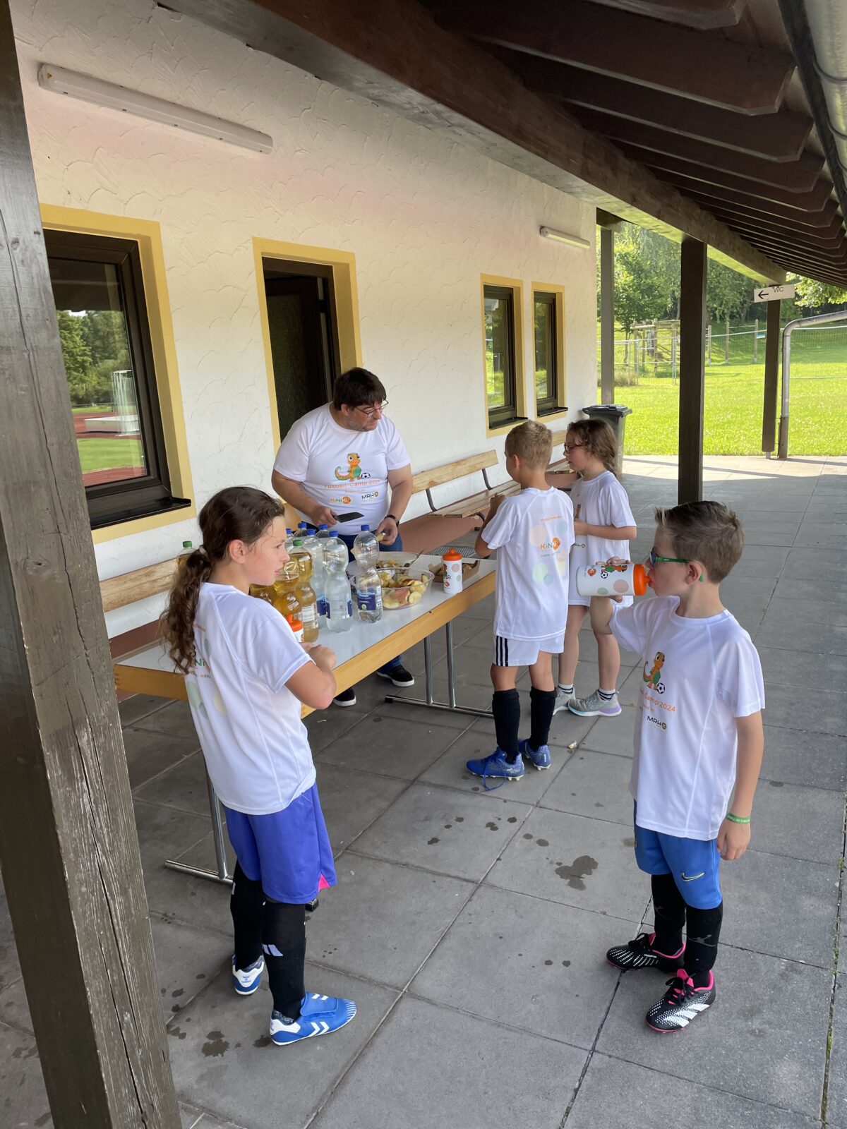 Eine Gruppe von Kindern in weißen T-Shirts versammelt sich um einen Holztisch unter einem überdachten Unterstand. Sie unterhalten sich und greifen nach Getränken und Snacks, die auf dem Tisch bereitstehen. Einige Kinder halten Plastikbecher in den Händen, während andere aufmerksam zuhören. Der Ort wirkt wie ein Sport- oder Freizeitgelände mit natürlicher Umgebung im Hintergrund.