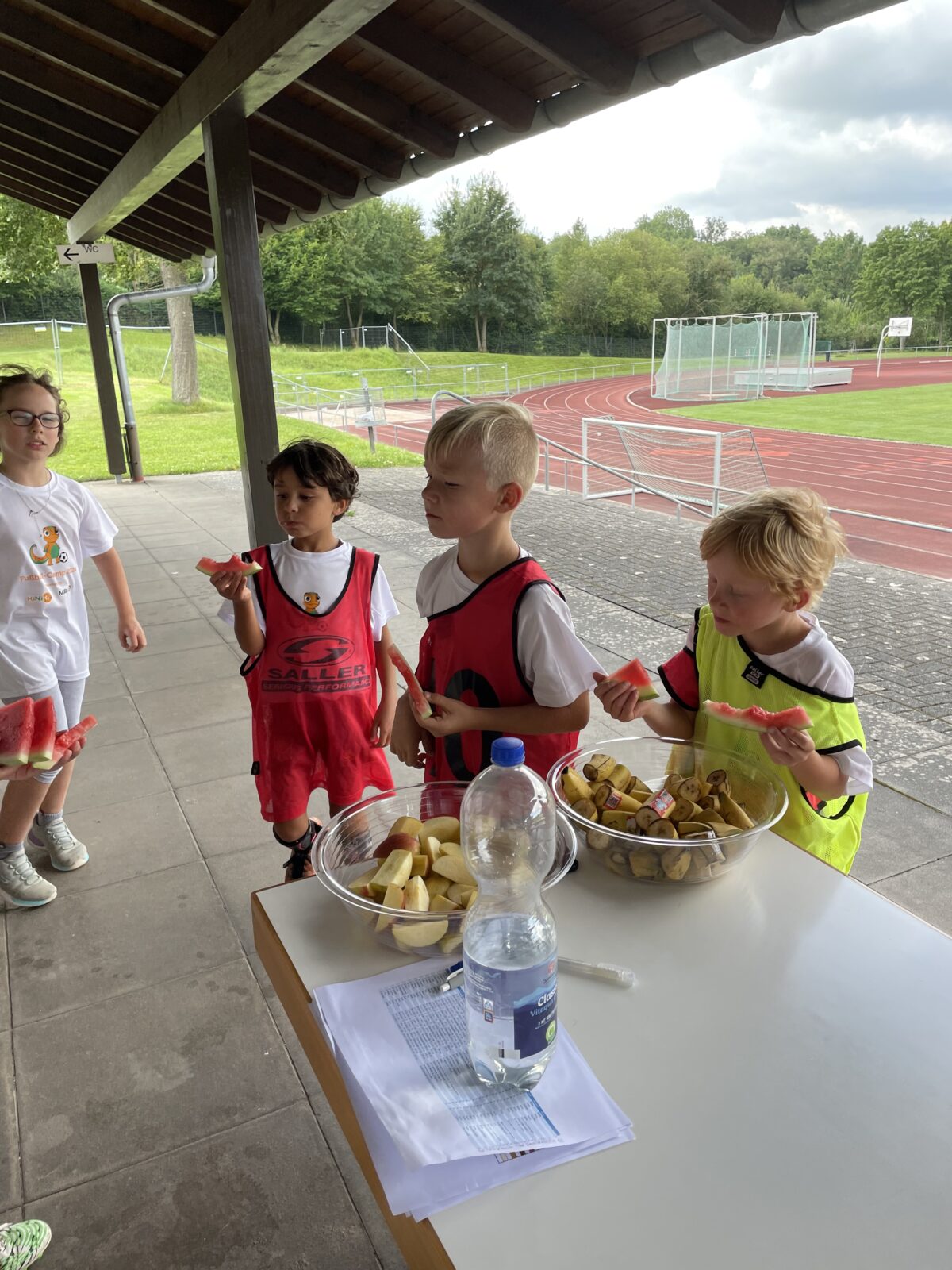 4 Kinder stehen an einem Tisch. Sie tragen Fußballklamotten. Auf dem Tisch steht eine Schale mit Äpfeln und eine mit Bananen. Ein Kind hat zwei Melonen Scheiben in der Hand. Sie stehen unter einem Vordach und im Hintergrund ist ein Sportplatz zu sehen.