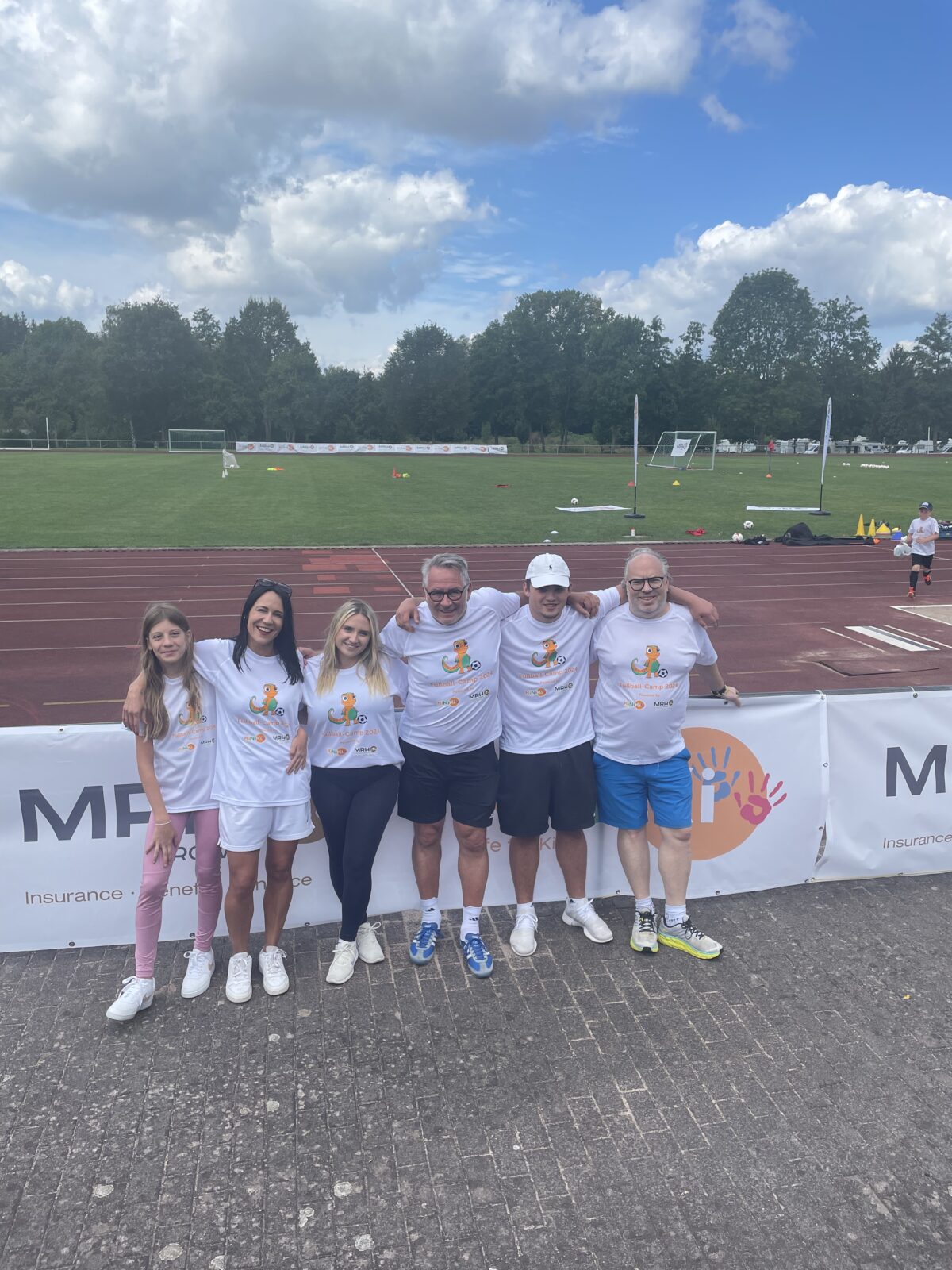 3 Frauen und 3 Männer stehen nebeneinander vor einem Zaun. Sie tragen weiße T-Shirts mit dem KiNiKI Gecko drauf. Der Zaun ist mit Bannern behangen die das KiNiKi Logo und MRH Trowe Logo zeigen Im Hintergrund sieht man einen Sportplatz und ein Fußballfeld.