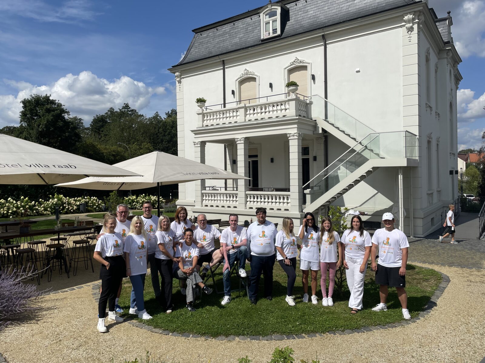 Bei sonnigem Wetter steht eine Gruppe Erwachsener steht vor einem Gebäude. Sie tragen alle weiße T-Shirts mit dem KiNiKi Gecko drauf und stehen aufgereiht nebeneinander.
