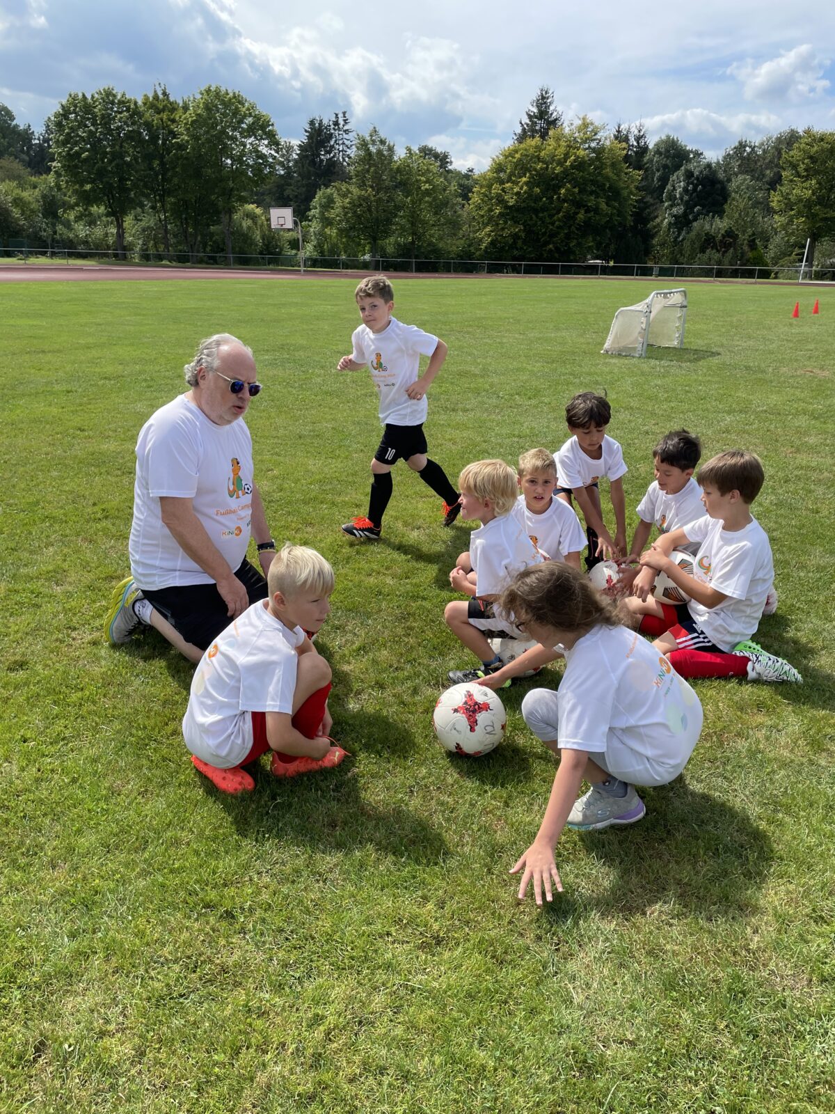 Eine Gruppe Kinder kniet auf dem Boden vor einem Mann auf einer Wiese. Sie tragen alle weiße T-Shirts mit dem KiNiKi Gecko drauf und tragen Fußballklamotten. Im Hintergrund sind Bäume und der blaue Himmel zu sehen.