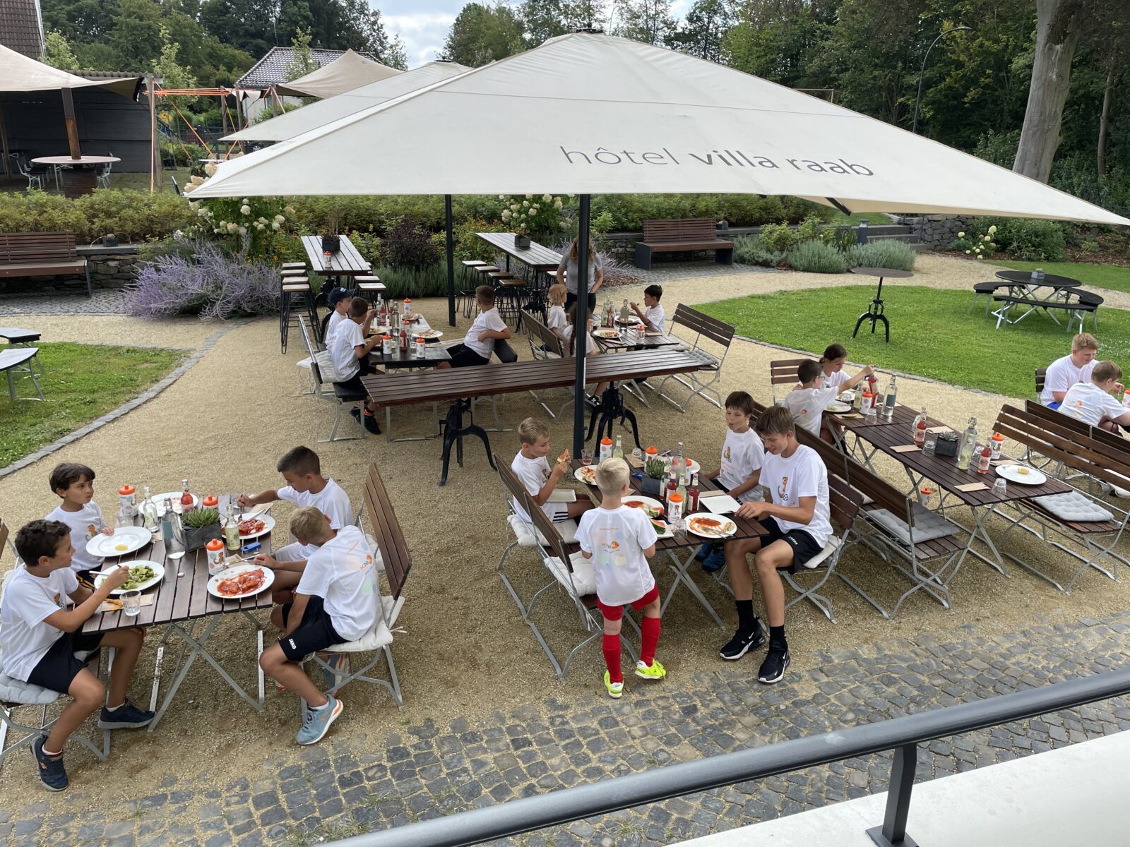 Mehrere Kinder sitzen gemeinsam mit Erwachsenen an Tischen unter einem großen weißen Sonnenschirm mit der Aufschrift „Hotel Villa Raab“. Sie unterhalten sich, essen oder trinken etwas und genießen eine Pause im Freien. Im Hintergrund sieht man eine Terrasse mit weiteren Sitzmöglichkeiten und Pflanzen, die für eine entspannte Atmosphäre sorgen.