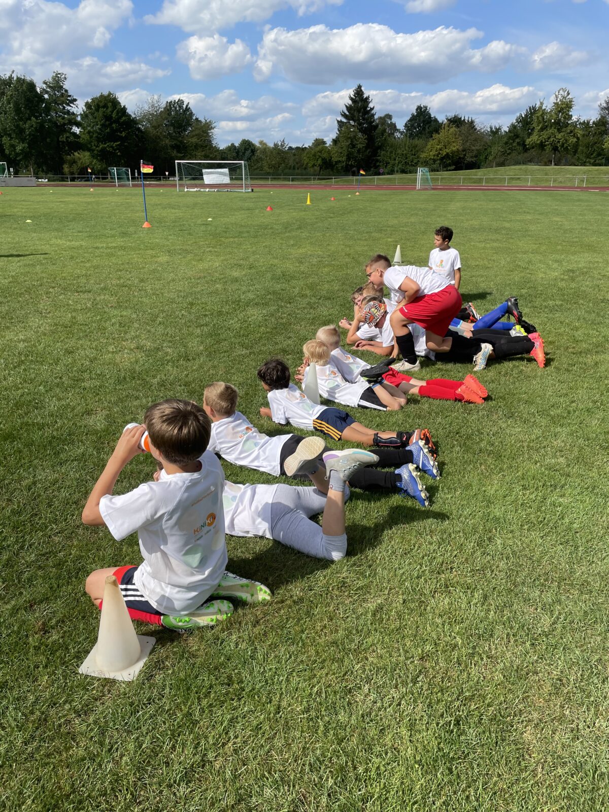 Eine Gruppe Kinder liegen nebeneinander auf einer Wiese und schauen in die Ferne. Sie tragen weiße T-Shirts und Fußballklamotten. Im Hintergrund sieht man einige Bäume und den blauen Himmel mit ein paar Wolken.