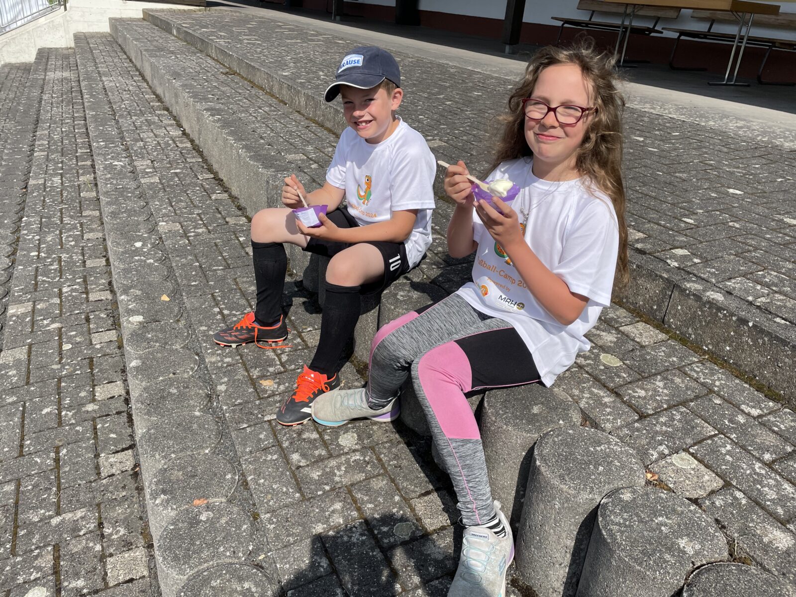 Zwei Kinder, ein Junge und ein Mädchen sitzen auf einer Treppe. Sie tragen beide Fußballklamotten und halten einen Becher mit Eis in der Hand und lächeln freundlich in die Kamera.