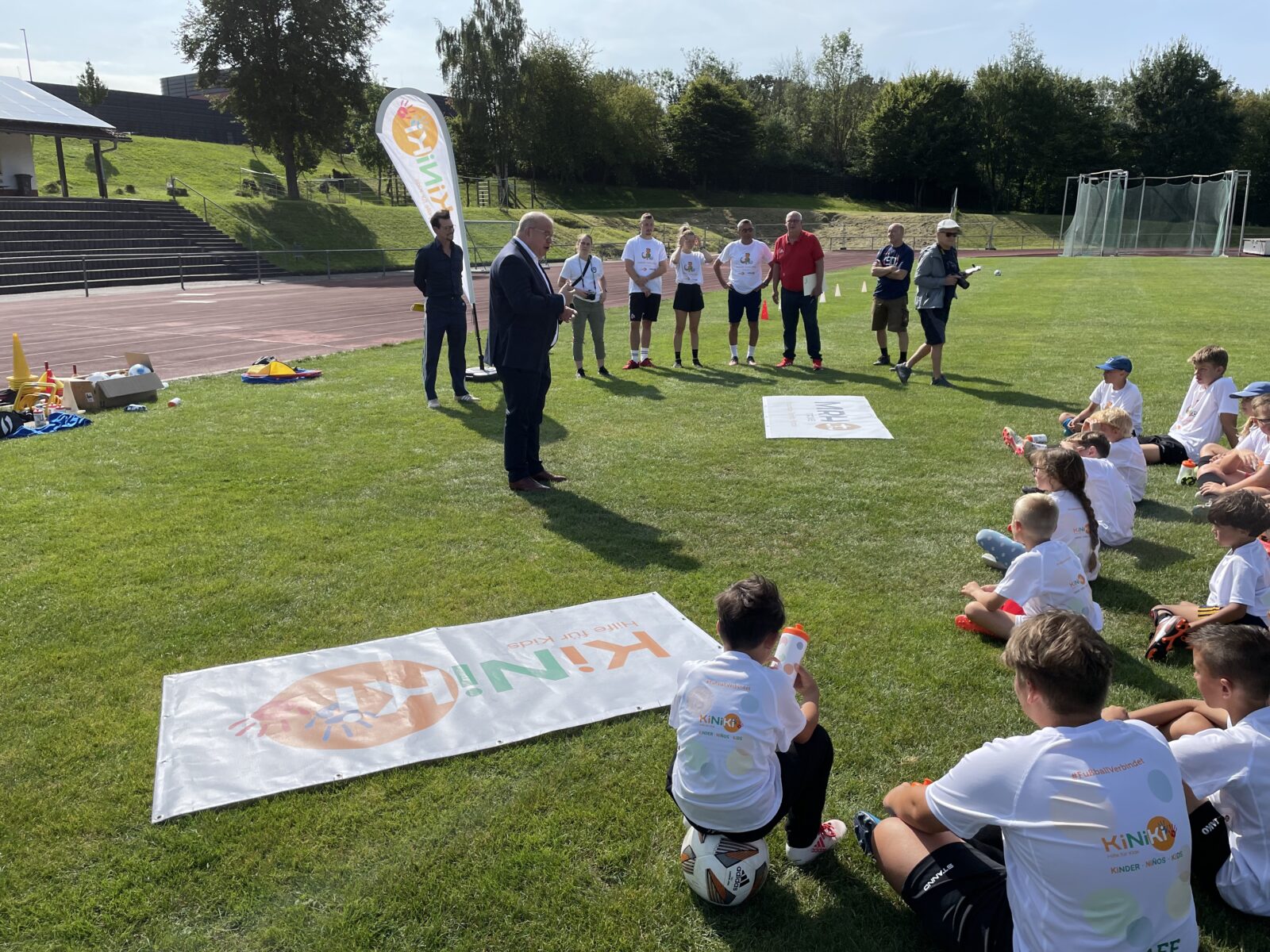 Eine Gruppe Kinder sitzt auf einem Fußballfeld und sie tragen weiße T-Shirts, mit dem KiNiKi Logo drauf. Vor den Kindern stehen Erwachsene. Ein Erwachsener trägt einen Anzug und hält eine Ansprache.