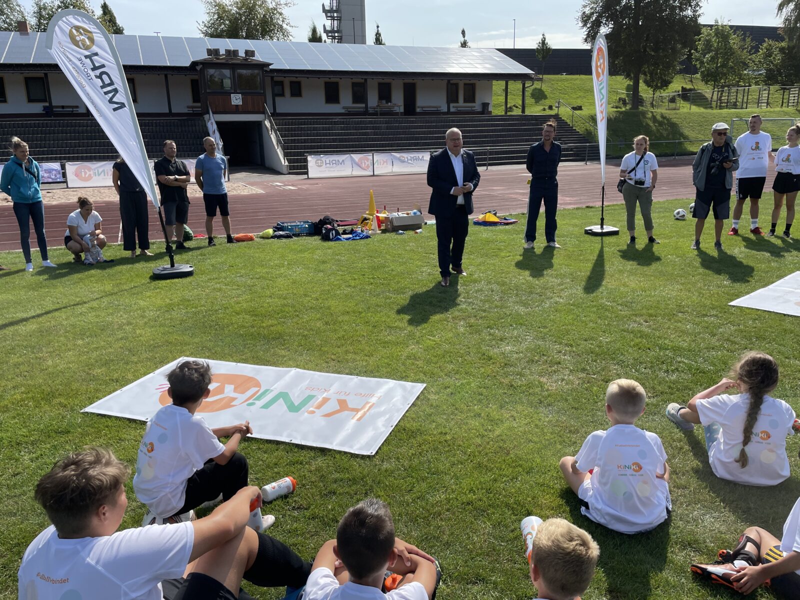 Eine Gruppe Kinder sitzt auf einem Fußballfeld und sie tragen weiße T-Shirts, mit dem KiNiKi Logo drauf. Vor den Kindern stehen Erwachsene. Ein Erwachsener trägt einen Anzug und hält eine Ansprache.