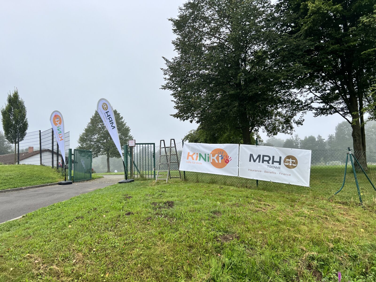 Das Bild zeigt eine Wiese und einen Zaun. An dem Zaun hängen zwei Banner, auf dem einen ist das KiNiKi Logo und auf dem anderen das MRH Trowe Logo. Es ist bewölkt.