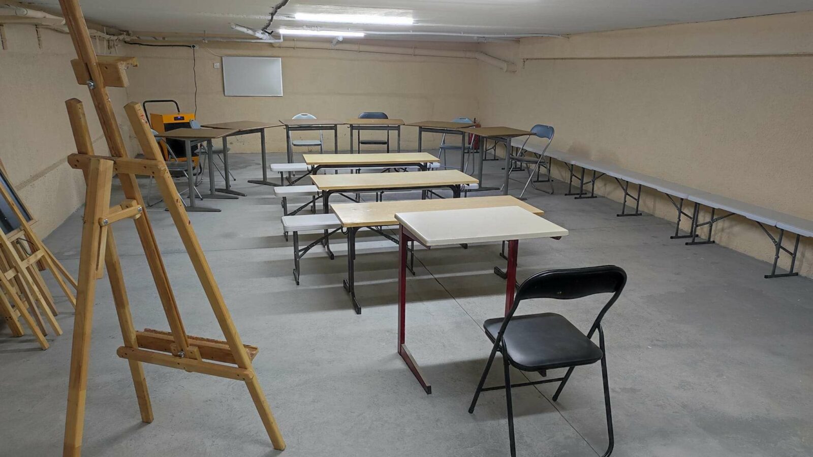Zu sehen ist ein düsterer Raum im Keller eines Gebäudes. Der Raum soll als Schulraum für Kinder genutzt werden. Es stehen einzelne Tische mit Stühlen verteilt im Raum.