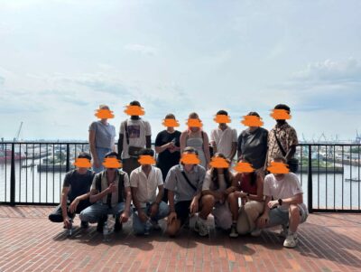 Das Bild zeigt eine Gruppe von 14 Personen, die draußen vor einer Brüstung posiert, mit einem Wasserhafen oder einer Küstenlandschaft im Hintergrund. Einige Personen sitzen auf dem Boden, während andere stehen. Die Kleidung ist sommerlich und die Atmosphäre wirkt entspannt, als ob sie sich auf einem Ausflug oder einer gemeinsamen Reise befinden. Der Himmel ist leicht bewölkt, aber das Wetter scheint angenehm.