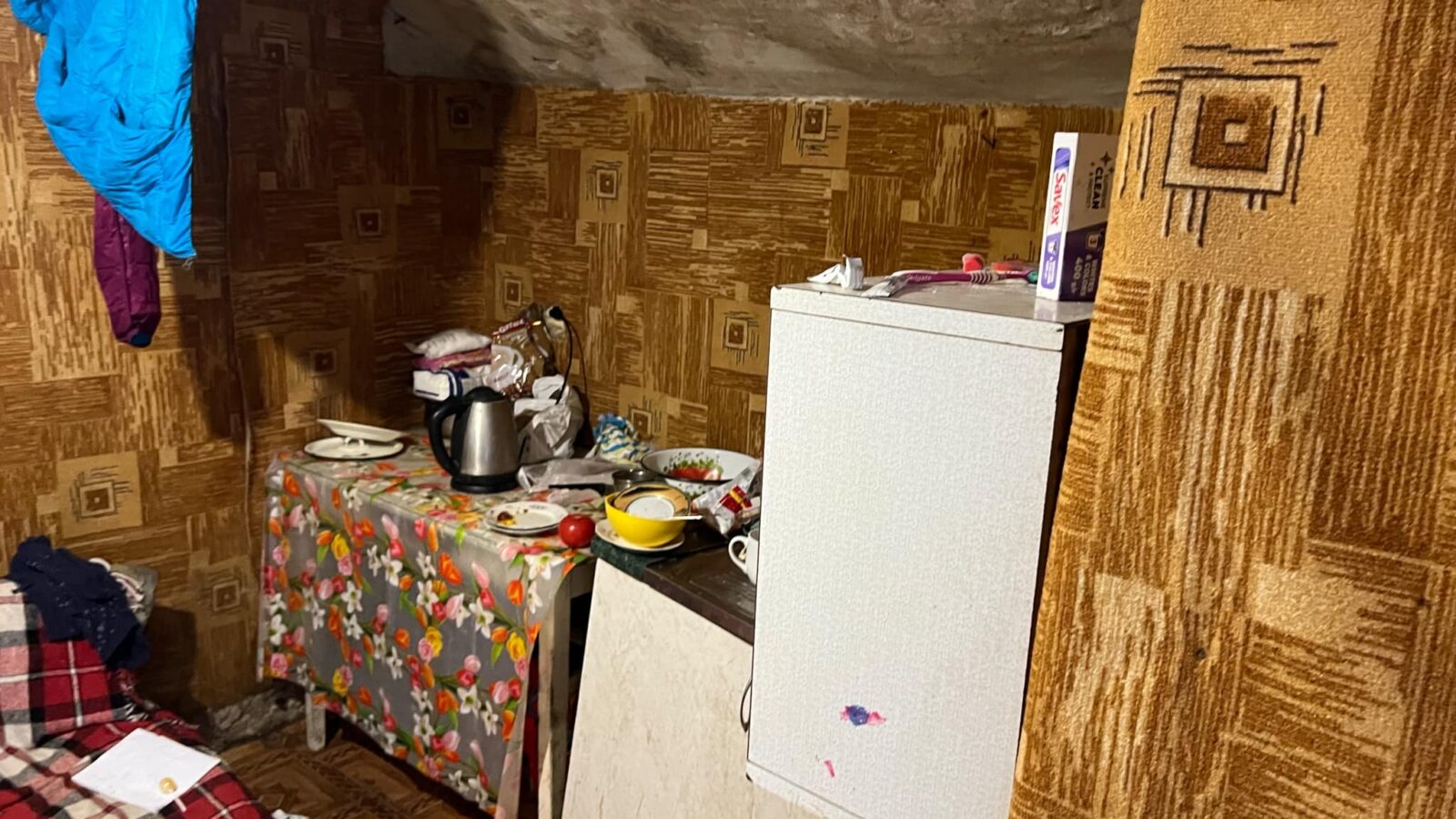 Ein kleiner Raum mit abgenutzter Tapete und einfacher Einrichtung. Auf einem kleinen Tisch mit geblümter Tischdecke stehen Teller, eine Schüssel, eine Teekanne und Lebensmittel. Daneben steht ein kleiner Kühlschrank, auf dem einige Hygieneartikel liegen. Die Decke ist schräg und verleiht dem Raum eine enge Atmosphäre.