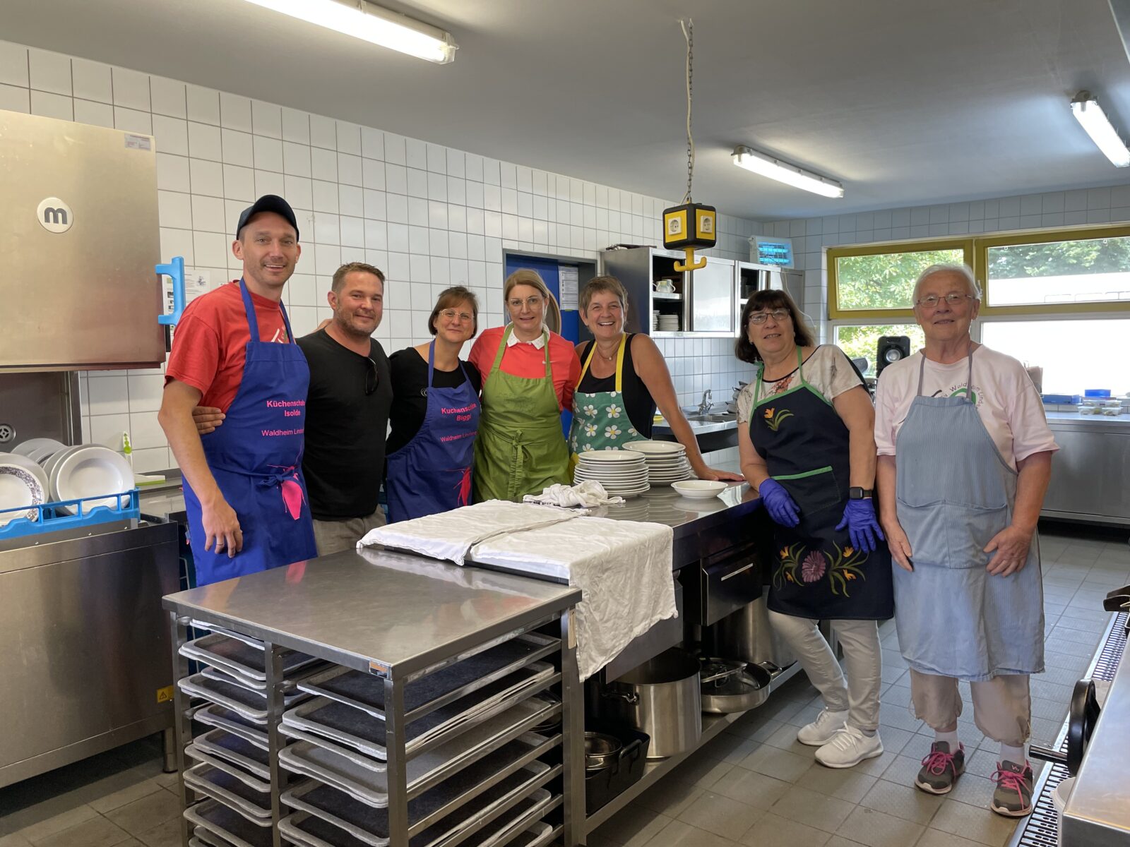 7 Personen stehen in einer Küche zusammen, tragen bunte Schürzen und lächeln freundlich in die Kamera. Vor ihnen stehen saubere Teller.