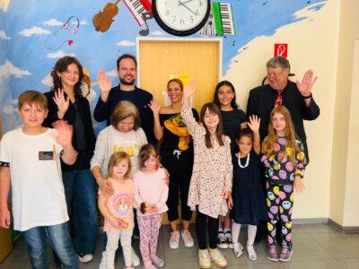 Das Bild zeigt eine Gruppe Menschen bestehend aus Kindern und Erwachsenen. Sie stehen vor einer Wand, auf der Musikinstrumente sind. Sie schauen alle lächelnd und winken mit der rechten Hand in die Kamera.