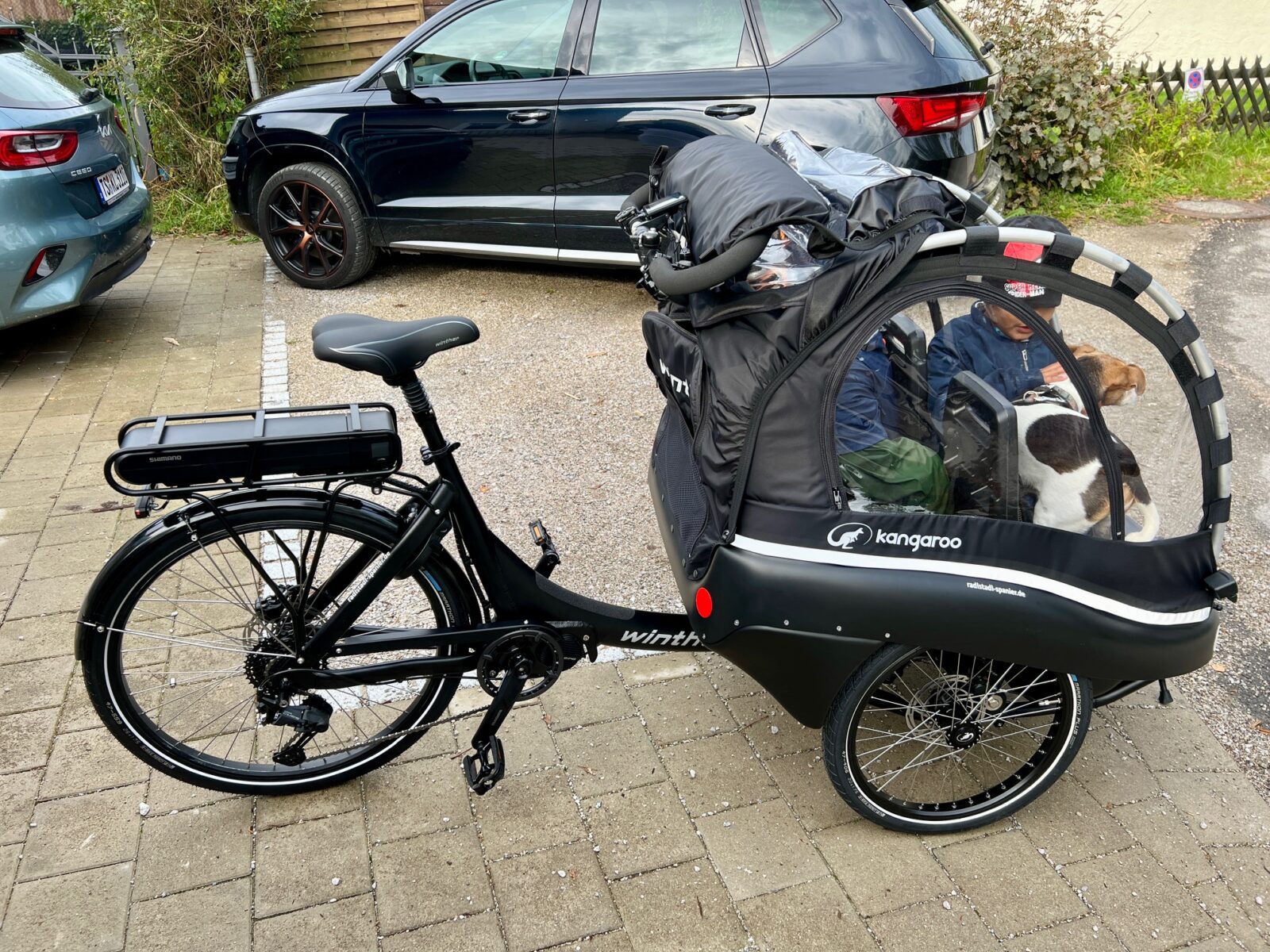 Das Bild zeigt ein schwarzes Lastenfahrrad der Marke "Winther" mit einem geschlossenen Anhänger der Serie "Kangaroo". Im Anhänger sitzen zwei Kinder und ein kleiner braun-weißer Hund. Die Kinder sind durch ein großes, transparentes Fenster zu sehen. Das Fahrrad ist mit einem Shimano-Elektroantrieb ausgestattet, und der Akku ist am Gepäckträger befestigt. Im Hintergrund sind geparkte Autos und etwas Grünfläche zu erkennen. Das Fahrrad steht auf einem gepflasterten Parkplatz.