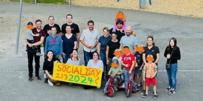 Ein Gruppenfoto mit allen Helfenden und den Kindern. Sie stehen nah beieinander und lächeln in die Kamera. In der vorderen Reihe wird das bemalte Plakat gehalten. Im Hintergrund sieht man recht die Schaukel und den Sandkasten. Links sieht man die frisch gestrichenen Beete.