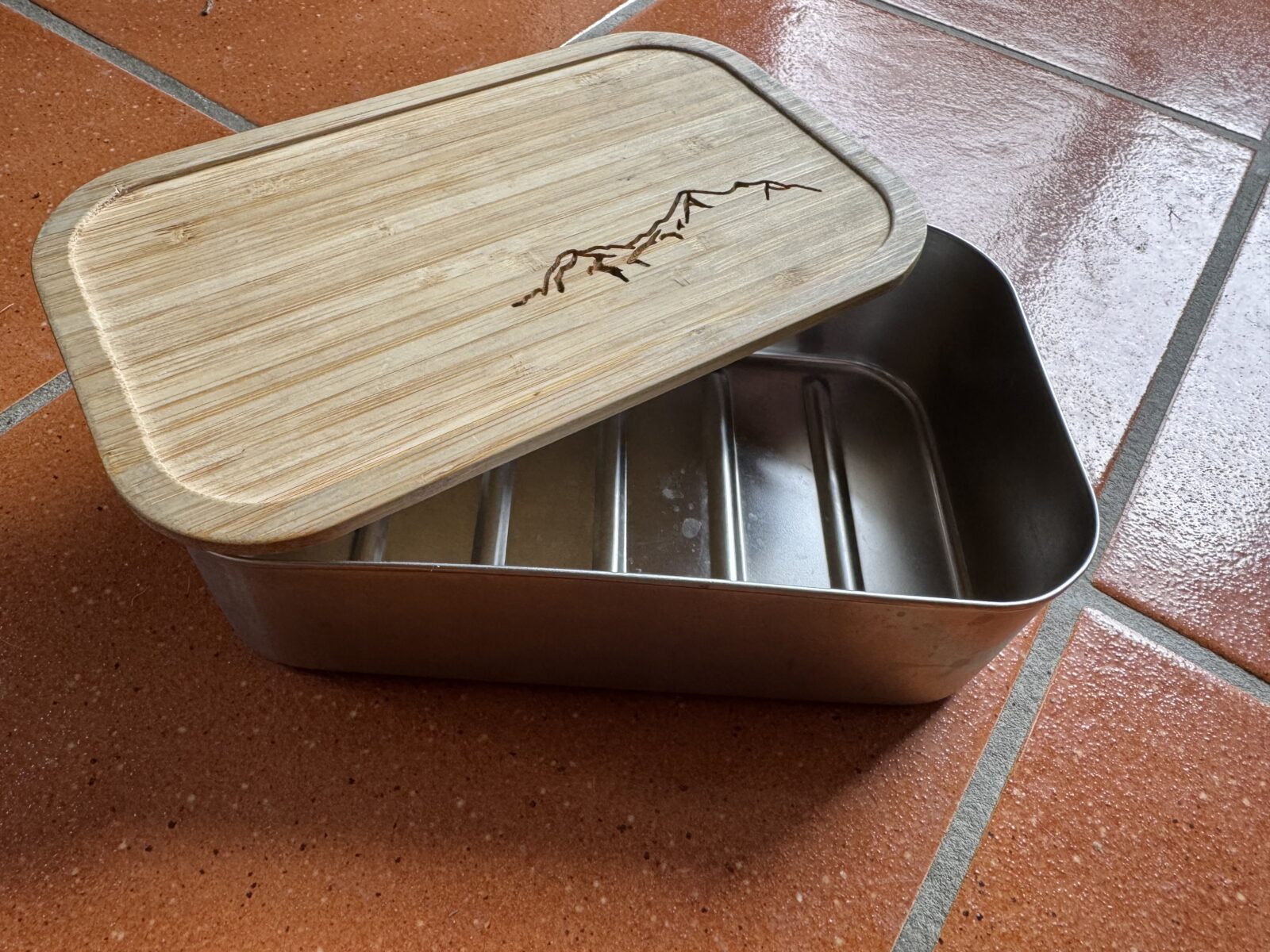 Eine wiederverwendbare Edelstahl-Brotzeitbox mit einem Bambusdeckel, der mit einem Bergmotiv graviert ist. Die Box liegt auf einem terrakottafarbenen Boden.