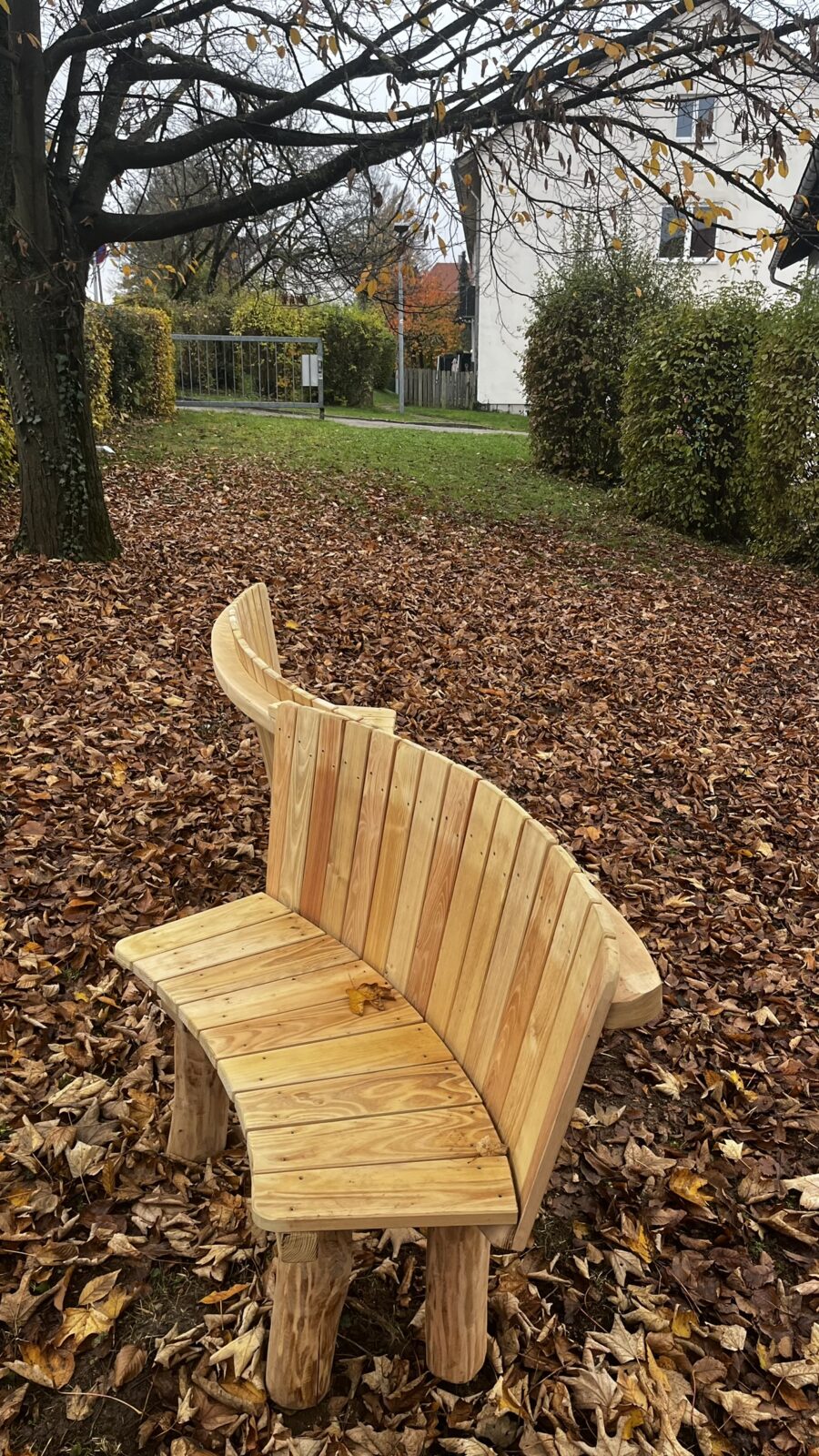 Das Bild zeigt zwei Holzbänke, die aneinander stehen und so gestellt sind, dass sie eine Art Welle Formen. Sie stehen auf Laub, vermutlich in einem Park. Im Hintergrund sieht man einen Baum, der fast all seine Blätter verloren hat und ein Reihenhaus.