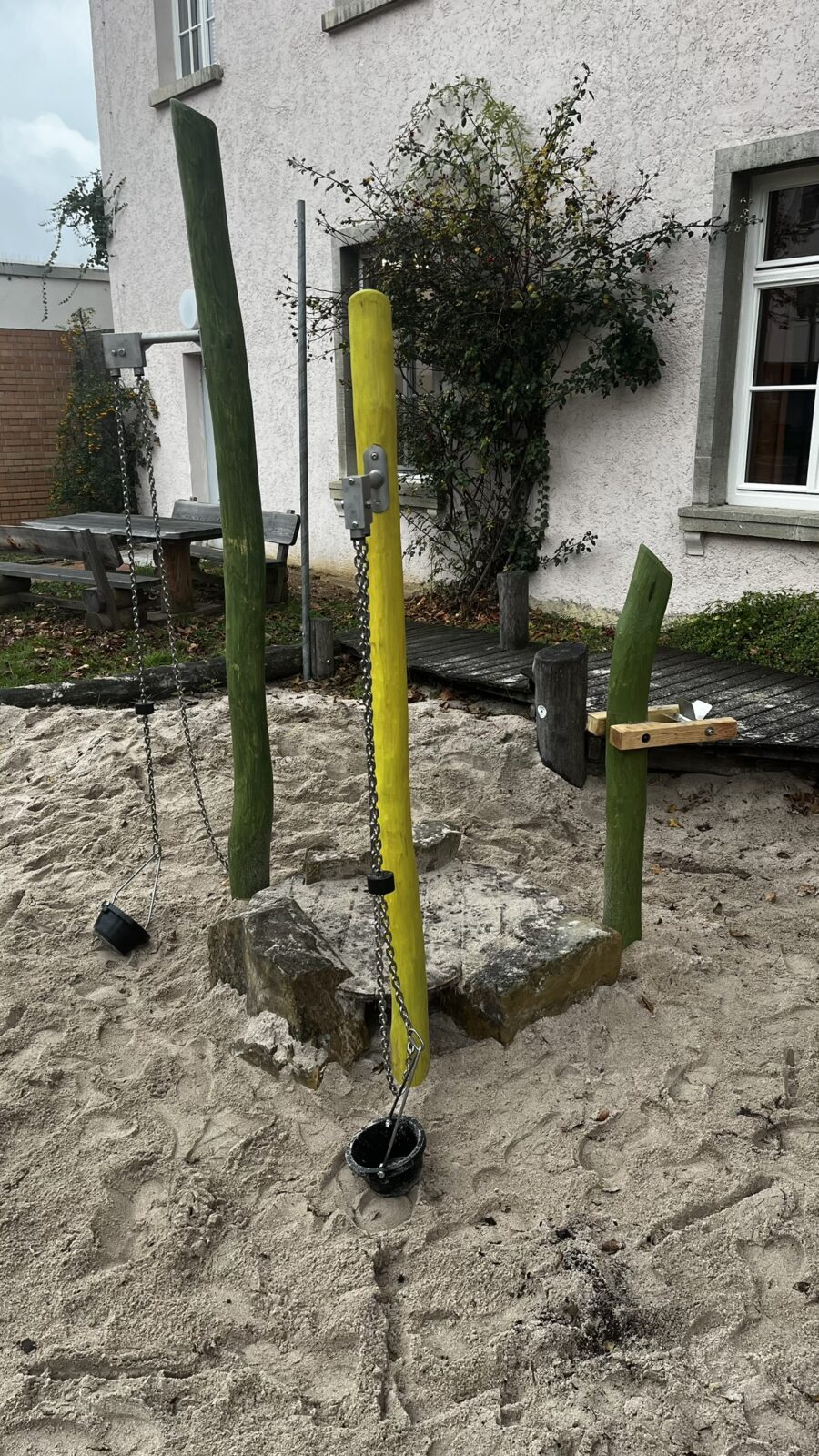 Das Bild zeigt einen Sandkasten mit 3 bunten Holzpfälern. Man kann mit dieser Konstruktion den Sand im Eimer hoch und runter ziehen.