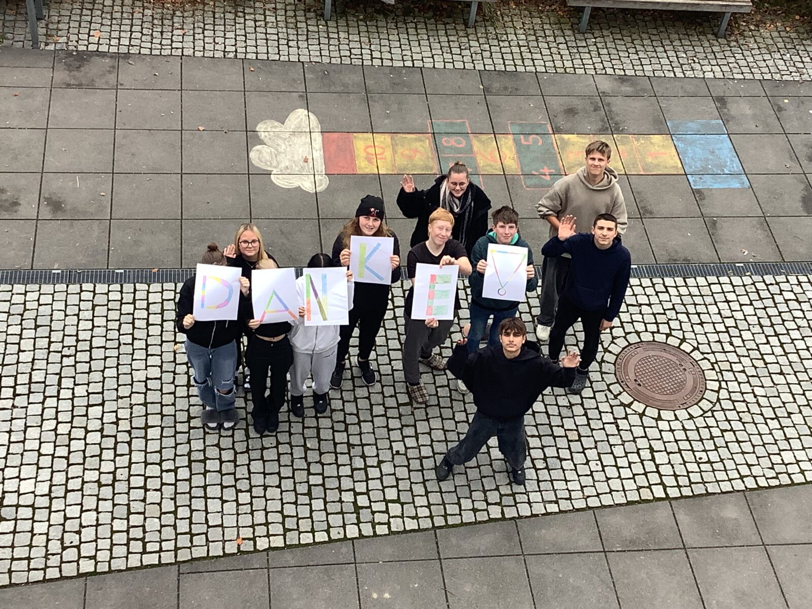 Das Bild zeigt eine Gruppe von Personen, die auf einem gepflasterten Platz stehen und nach oben blicken. Sie halten Blätter in den Händen, die zusammen das Wort "DANKE!" bilden. Einige Personen winken, und es ist farbige Straßenkreide auf dem Boden zu sehen, darunter ein Hüpfspiel mit Zahlen. Die Stimmung wirkt freundlich und dankbar.