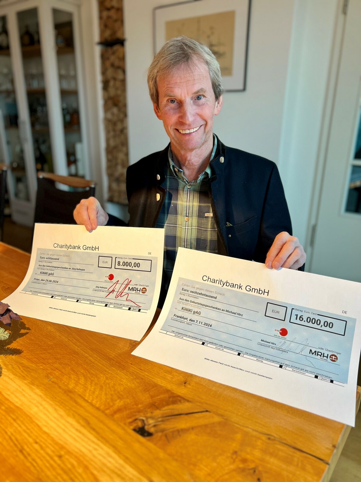 Ein Mann sitzt lächelnd an einem Holztisch und präsentiert zwei große symbolische Schecks: einen über 8.000 Euro und einen über 16.000 Euro. Beide Spenden stammen aus Geburtstagsaktionen und sind für KiNiKi bestimmt.