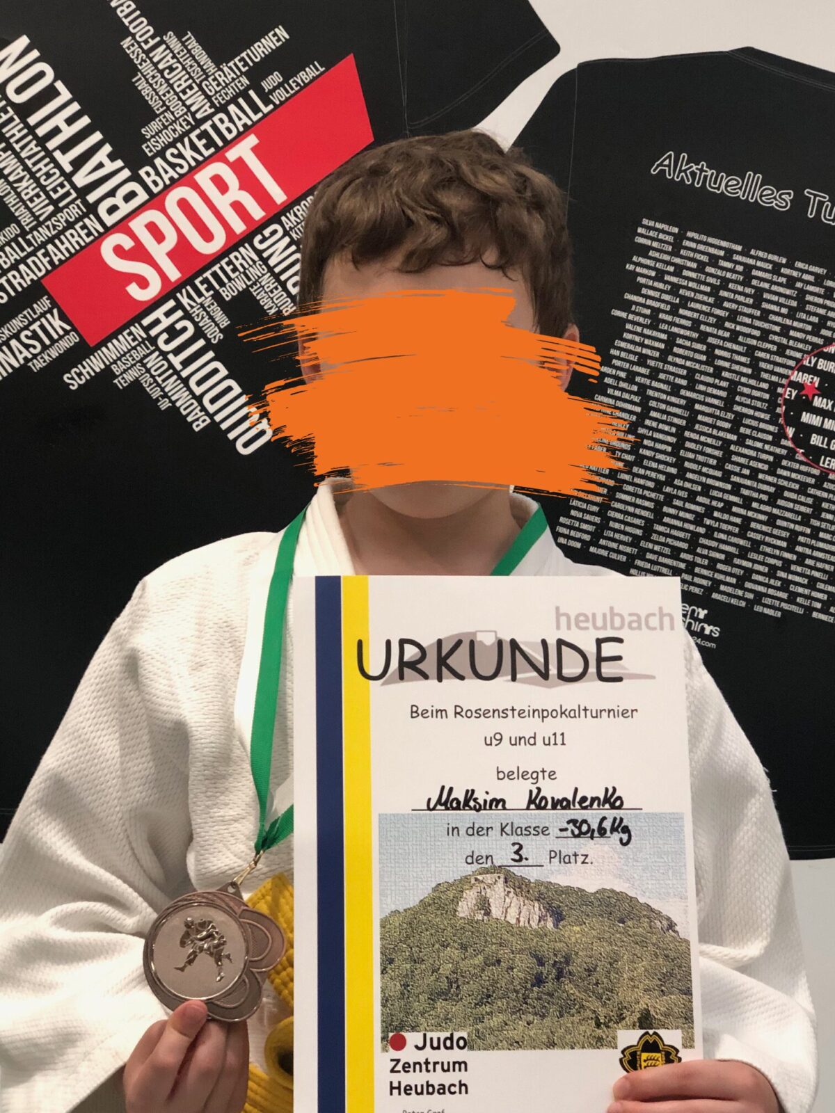 Ein Junge mit Judo An zug hält stolz seine Medaille in der einen und seine Urkunde in der anderen Hand. Hinter ihm hängen zwei Sport T-Shirts.