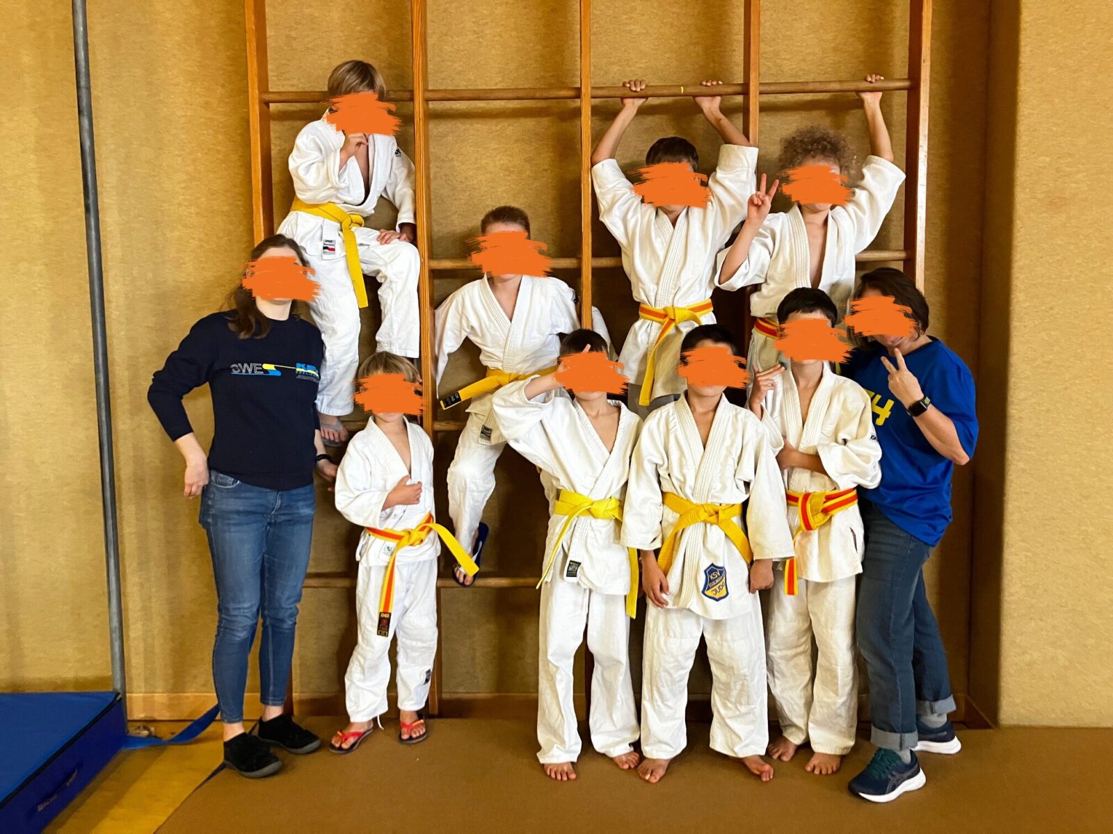 Das Bild zeigt insgesamt 8 Jungs und zwei Frauen. Sie stehen als Gruppe vor einer Holz-Kletterwand. 4 Jungs hängen an an der Kletterwand. Sie lächeln alle freundlich in die Kamera und manche zeigen mit ihren Fingern "Peace".