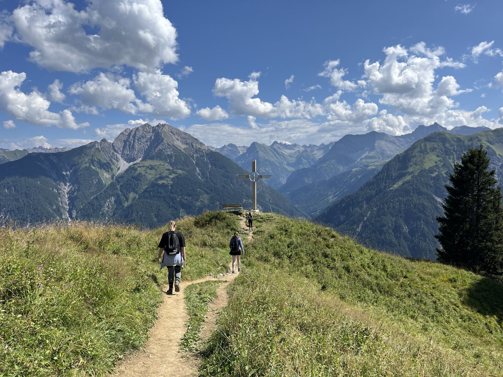 Ein schmaler Wanderpfad führt zwei Personen entlang einer grünen Bergwiese hin zu einem Gipfelkreuz. Im Hintergrund erstreckt sich eine beeindruckende Alpenlandschaft mit schroffen Berggipfeln und einem strahlend blauen Himmel mit weißen Wolken.