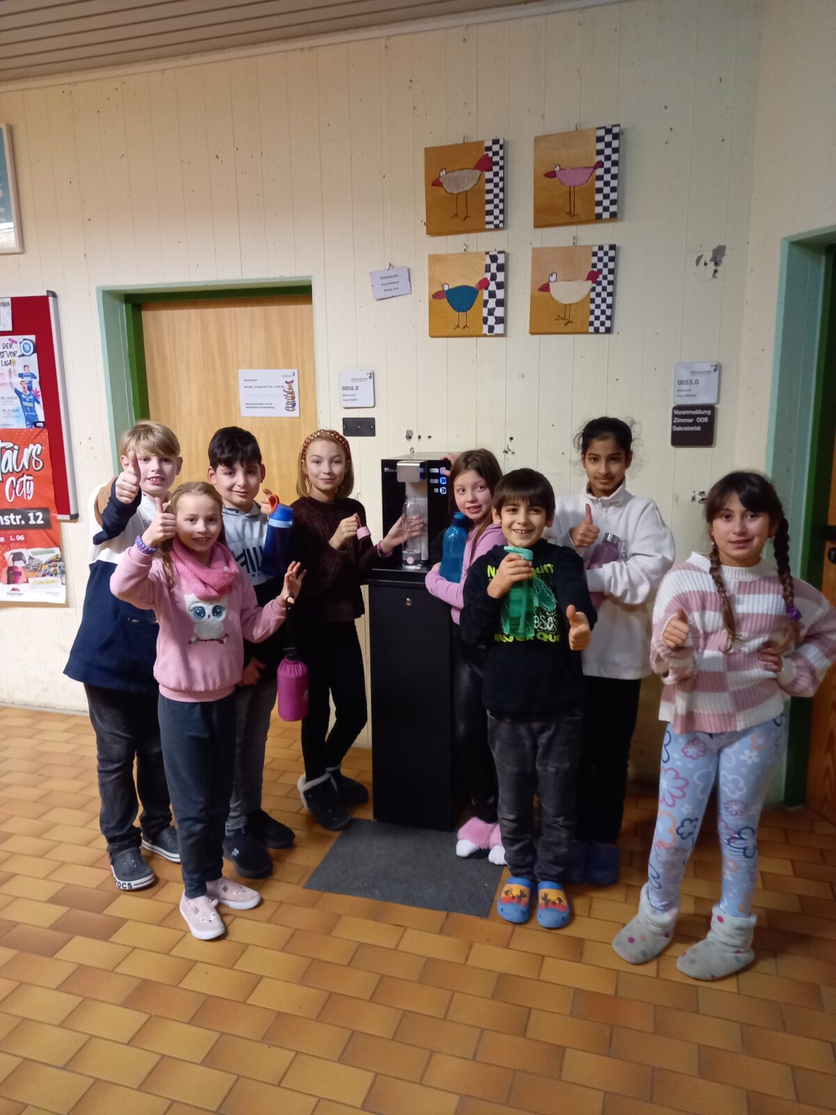 Eine Gruppe von acht Kindern steht vor einem Wasserspender in einem Schulflur. Die Kinder lächeln, zeigen Daumen hoch und halten teils Trinkflaschen. Sie tragen warme Kleidung und gemütliche Hausschuhe oder Socken. Im Hintergrund sind bunte Wandbilder mit Vogelmotiven sowie Türen zu sehen. Der Boden ist mit braunen Fliesen ausgelegt.