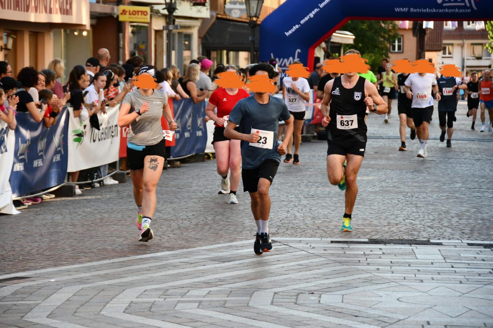 Eine Gruppe von Läufern, darunter ein junger Teilnehmer, läuft während eines Stadtlaufs durch die Innenstadt. Die Zuschauer am Rand feuern die Läufer an, während die Teilnehmenden unter einem Start-/Zielbogen hindurchlaufen. Die Atmosphäre ist lebendig, mit Jubel und sportlichem Einsatz.