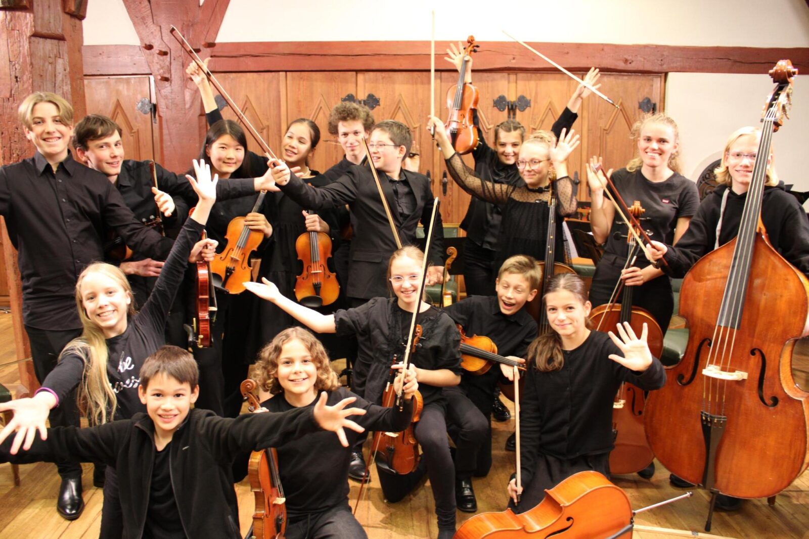 Eine Gruppe von Jugendlichen mit Musikinstrumenten wie Geigen, Cello und Kontrabass posiert fröhlich in einem Konzertsaal. Alle tragen schwarze Kleidung und strahlen Freude und Begeisterung aus, während sie ihre Instrumente hochhalten. Im Hintergrund sind Holzverkleidungen und eine warme, einladende Atmosphäre zu sehen.