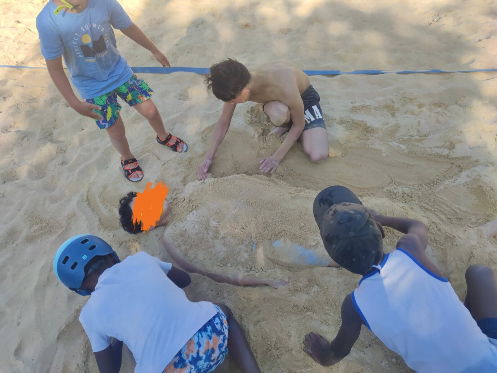 Eine Gruppe von Kindern spielt im Sand und hat einen Freund fast vollständig eingegraben, nur der Kopf schaut noch heraus. Die Kinder sind vertieft ins Spiel, lachen und genießen den sonnigen Tag. Die Szene strahlt Freude und Gemeinschaft aus.