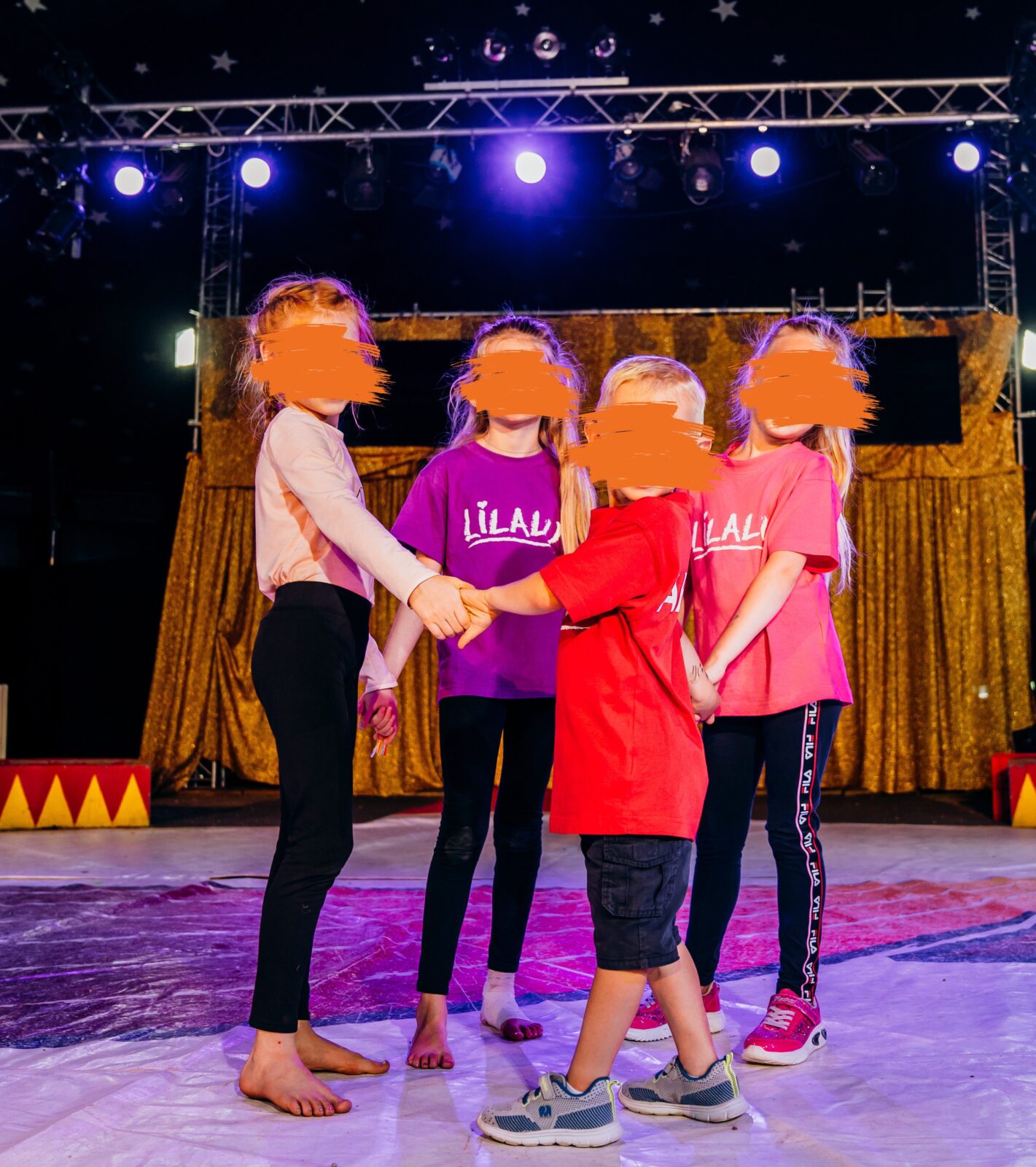 Das Bild zeigt 4 Kinder, 3 Mädchen und einen Jungen, die sich an der Handhalten und im Kreis stehen. Sie schauen alle lächelnd in die Kamera. Die Kinder tragen bunte T-Shirts mit der Aufschrift "Lilalu". Sie stehen auf einer Bühne und im Hintergrund sind ein goldfarbener Vorhang und Scheinwerfer zu sehen.