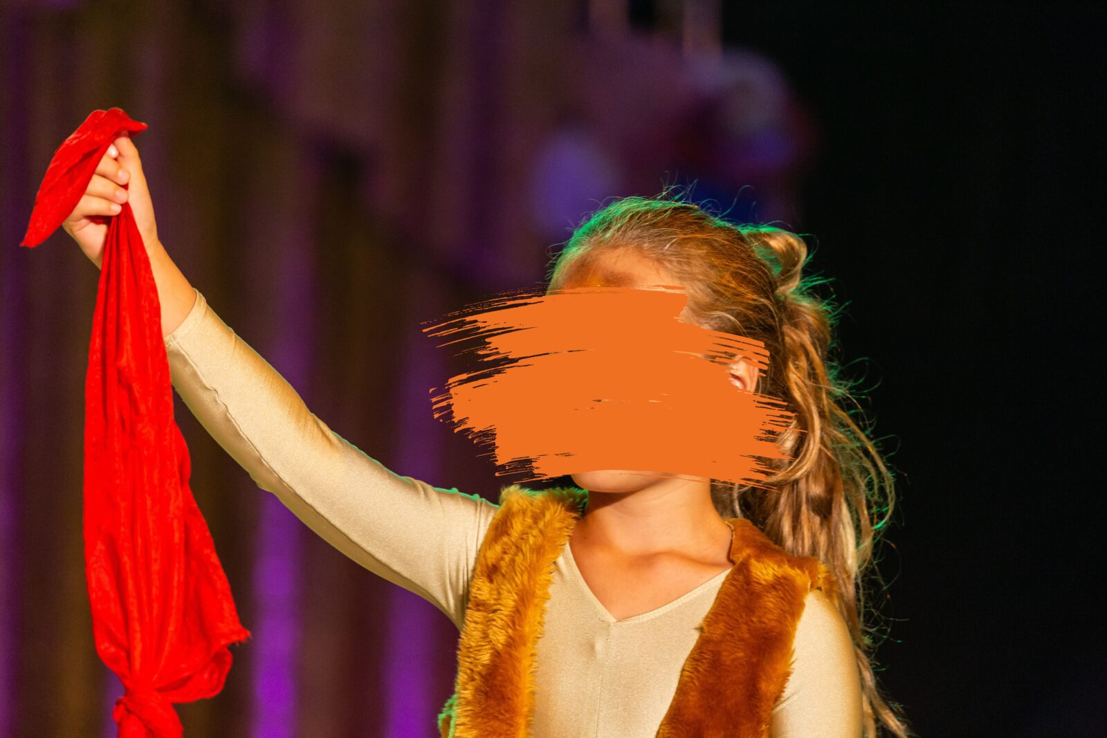 Ein kleines Mädchen ist wie ein Tiger geschminkt. Ihre Haare hat sie zu einem Pferdeschwanz gebunden und sie trägt eine Fellweste und darunter ein beigefarbenes Langarmshirt. Die eine Hand hat sie in der Luft und in der Hand hält sie ein rotes Tuch. Sie steht auf einer Bühne und der Hintergrund ist unscharf.