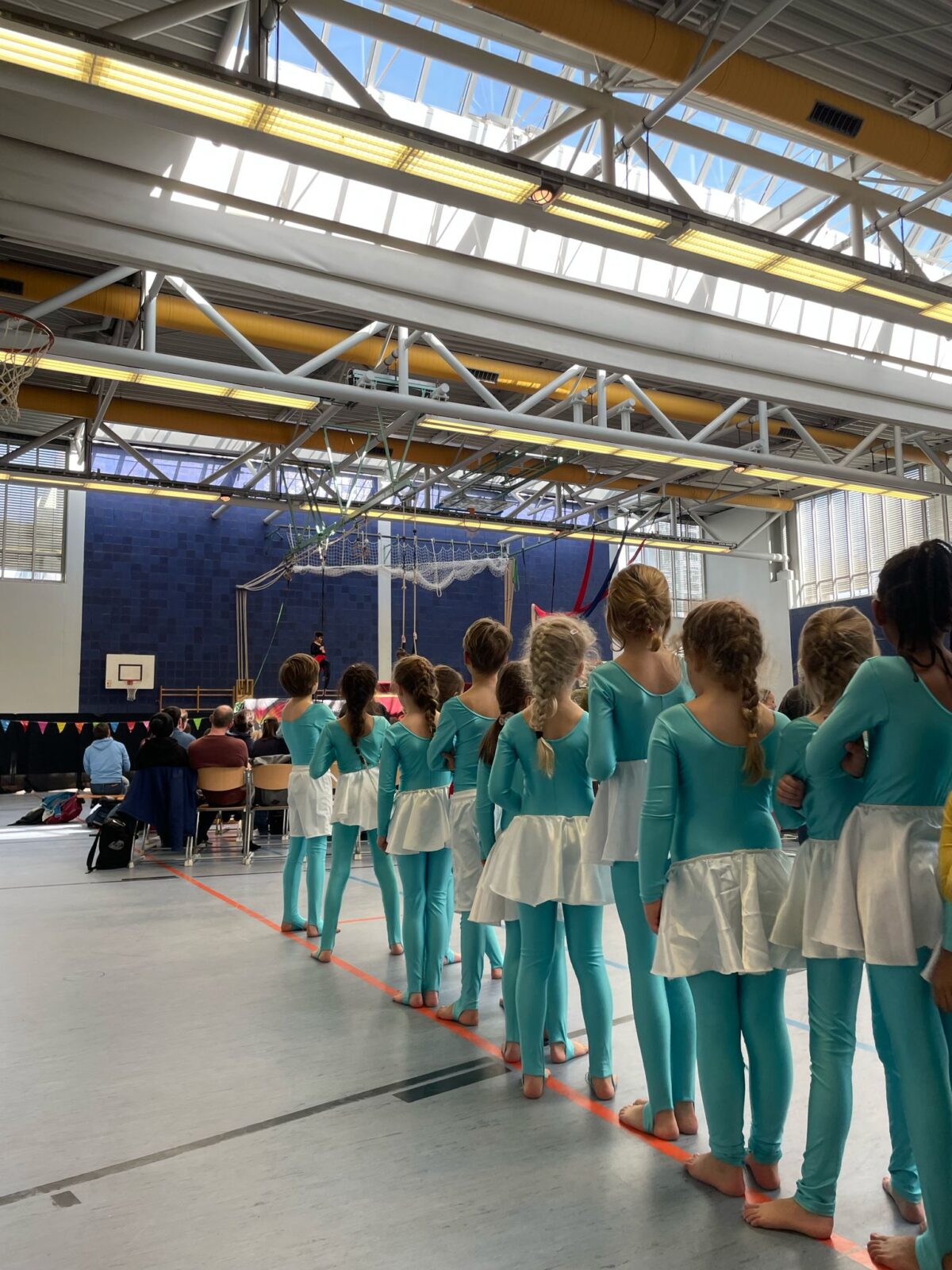 Eine Gruppe von jungen Mädchen steht barfuß in einer Turnhalle in einer Reihe. Sie tragen hellblaue Ganzkörperanzüge mit weißen Röckchen. Im Hintergrund sind Zuschauer auf Stühlen, eine Bühne und Turngeräte wie Seile und Netze zu sehen. Die Turnhalle hat ein hohes Glasdach mit Metallträgern.