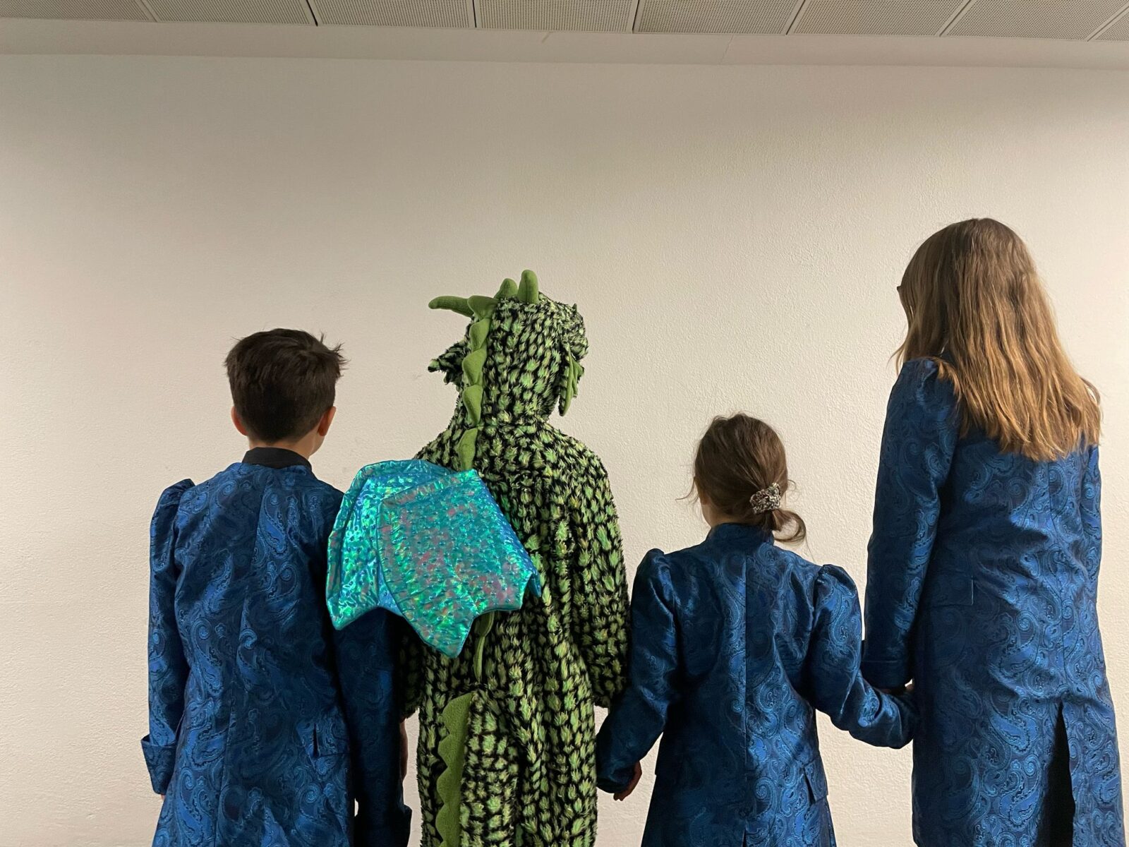 Vier Kinder stehen mit dem Rücken zur Kamera vor einer weißen Wand. Drei von ihnen tragen blaue, gemusterte Mäntel, während die vierte Person in der Mitte ein grünes, strukturiertes Drachenkostüm mit schimmernden blauen Flügeln trägt. Zwei der Kinder halten sich an den Händen.