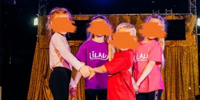 Das Bild zeigt 4 Kinder, 3 Mädchen und einen Jungen, die sich an der Handhalten und im Kreis stehen. Sie schauen alle lächelnd in die Kamera. Die Kinder tragen bunte T-Shirts mit der Aufschrift "Lilalu". Sie stehen auf einer Bühne und im Hintergrund sind ein goldfarbener Vorhang und Scheinwerfer zu sehen.