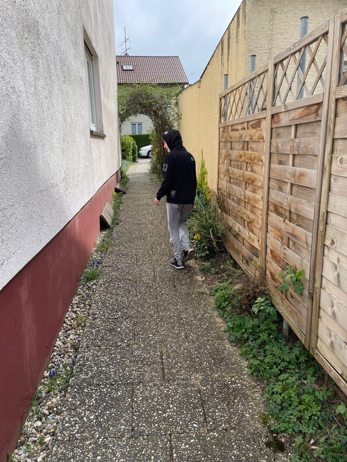 Das Bild zeigt eine Person, die hinter einem Haus lang läuft. Sie trägt einen schwarzen Kapuzenpulli und eine graue Jogginghose. Sie läuft Richtung eines Bogens, der aus Ästen geformt ist. Links neben der Person ist die Hauswand zu sehen und rechts von ihr ist ein Holzzaun.
