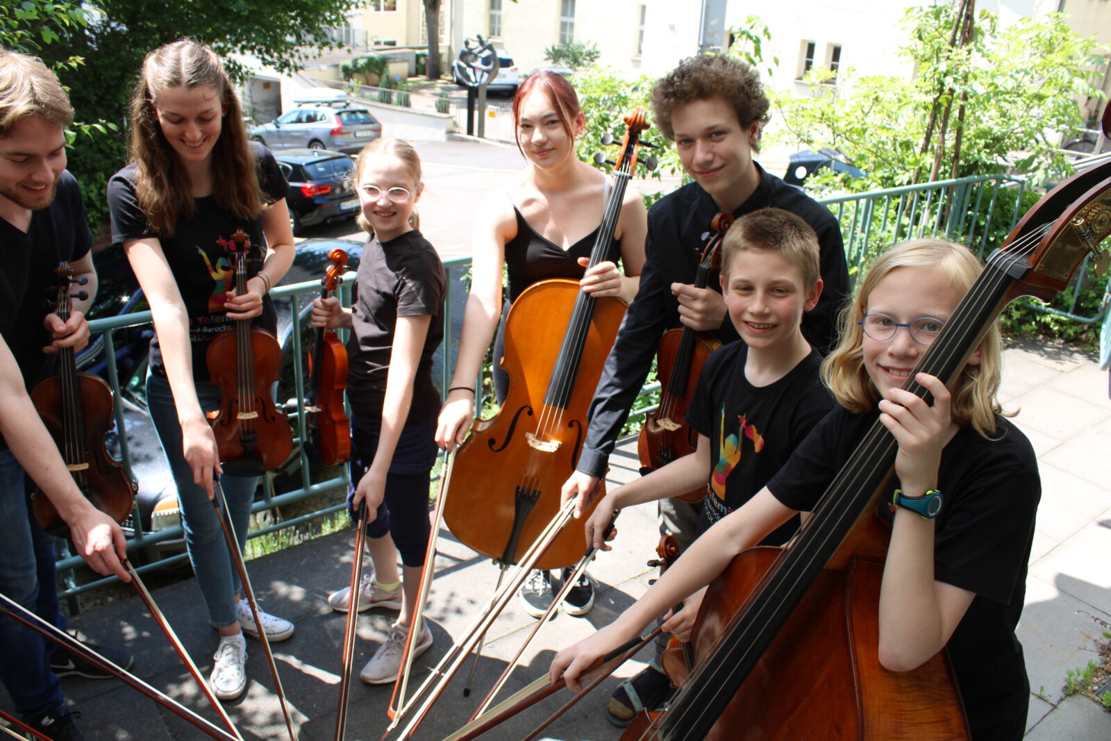 Eine Gruppe Kinder steht draußen auf einem Balkon in einem Kreis. In der einen Hand halten sie jeweils eine Geige und in der anderen einen Geigenbogen. Sie tragen alle Schwarze Klamotten und lächeln freundlich in die Kamera. Im Hintergrund sind Häuser und Bäume zu sehen.