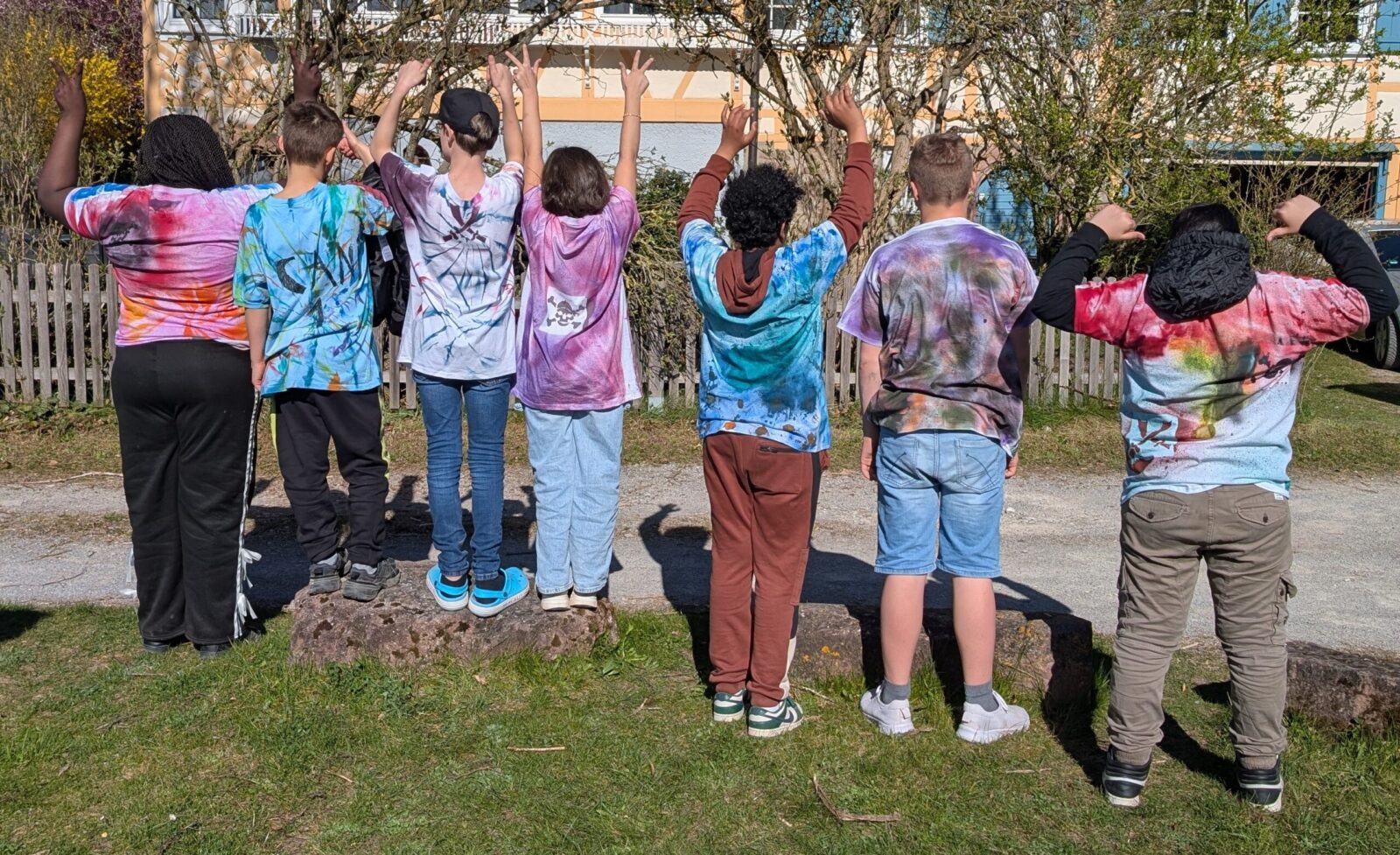 Eine Gruppe von sieben Kindern steht mit dem Rücken zur Kamera und hebt die Arme. Sie tragen bunte, selbstgestaltete T-Shirts und stehen im Freien auf Steinen, vor einem Zaun und Bäumen.