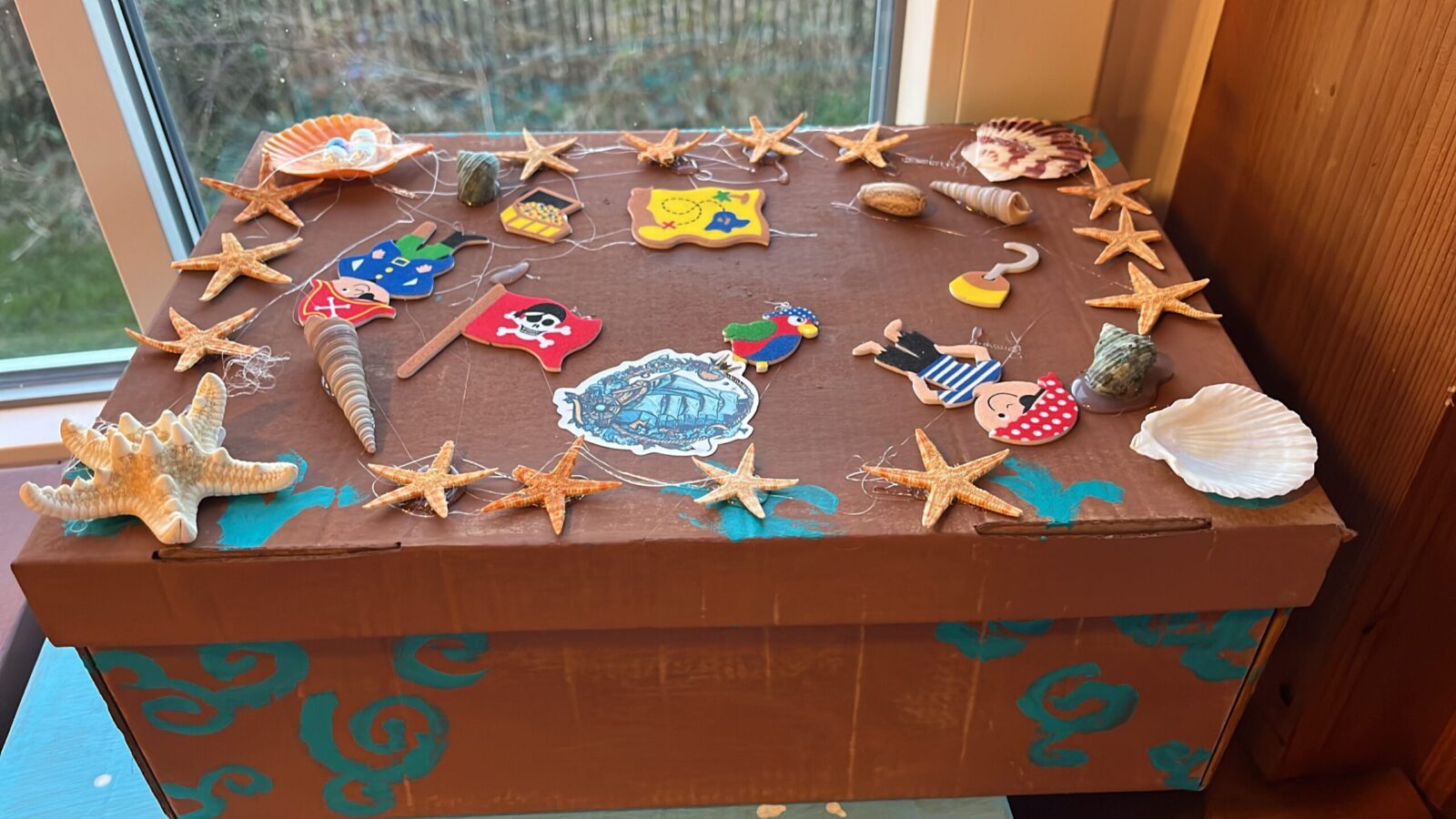Ein brauner Tisch mit verschiedenen Piraten-Dekorationen, darunter eine Schatzkarte, ein Piratenflagge, ein Papagei, eine Muschel, ein Haken, ein Pirat, ein Schatzkiste, mehrere Seesterne und ein Bild eines Schiffes. Im Hintergrund ist ein Fenster zu sehen.