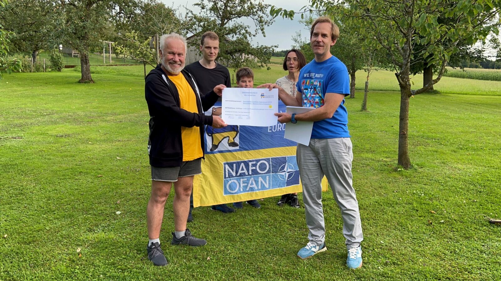 Eine Gruppe von Menschen steht in einem grünen Park und hält ein Zertifikat in den Händen. Im Hintergrund ist eine Flagge mit der Aufschrift "NAFO FAN" zu sehen, die auf Unterstützung für eine Organisation hinweist. Die Szene ist von Bäumen und einer Wiese umgeben, die eine friedliche und natürliche Atmosphäre schaffen.