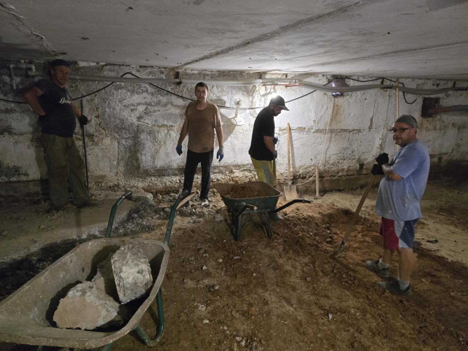 Vier Arbeiter stehen in einem Kellerraum und sind mit Bauarbeiten beschäftigt. Zwei Schubkarren, eine mit großen Steinen und eine leer, stehen im Vordergrund. Der Raum ist spärlich beleuchtet und die Wände sind roh und unverputzt. Der Boden scheint frisch ausgehoben zu sein und die Männer sind mit Werkzeugen wie Schaufeln und Spitzhacken ausgestattet.