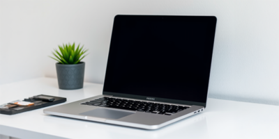 Moderner, silberner Laptop mit schwarzem Bildschirm auf einem weißen Schreibtisch neben einem kleinen Pflanzentopf und einem Grafiktablett, vor neutralem Hintergrund.