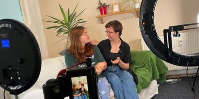 Zwei Personen sitzen auf einem weißen Sofa in einem gemütlichen Raum, während sie miteinander sprechen. Vor ihnen sind zwei große Ringlichter und eine Kamera auf einem Stativ positioniert, die offensichtlich eine Aufnahme machen. Im Hintergrund sind eine grüne Decke, Pflanzen und ein Regal mit Dekorationen zu sehen.